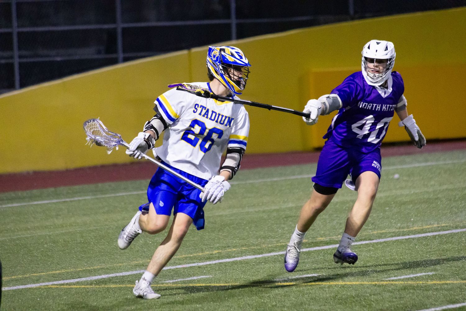 Two lacrosse players compete on a field at night, player #26 in blue and gold evading defender #40 in purple.
