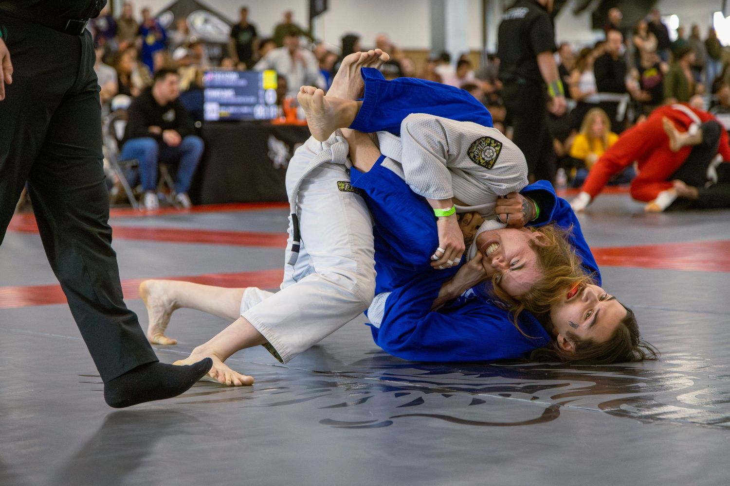 Two women competing in a Brazilian jiu-jitsu grappling match on a blue mat at an indoor tournament.