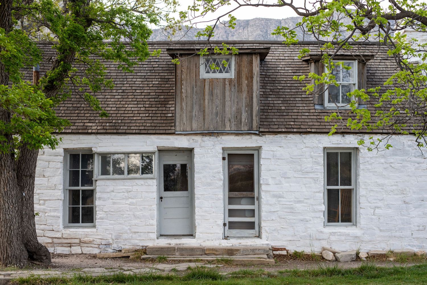 Historic white brick cottage with wooden dormer window, gray doors, and large tree, showcasing rustic frontier architecture.