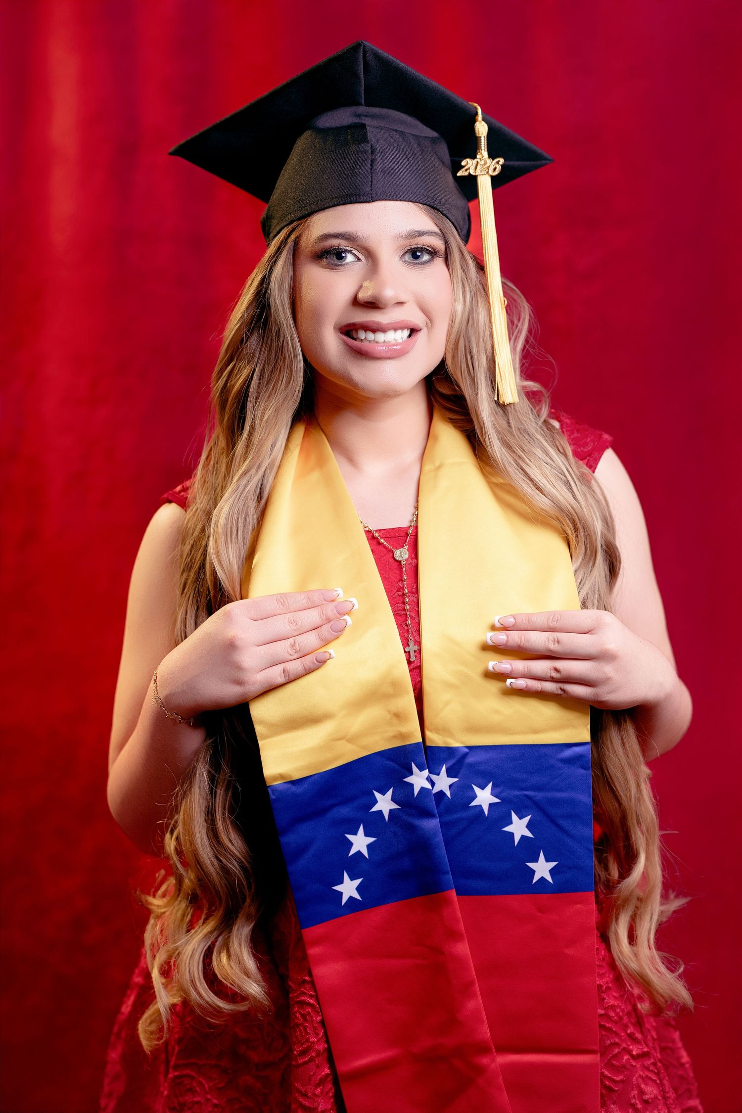 Young woman in graduation cap holds Venezuelan flag, smiling proudly against red background.
