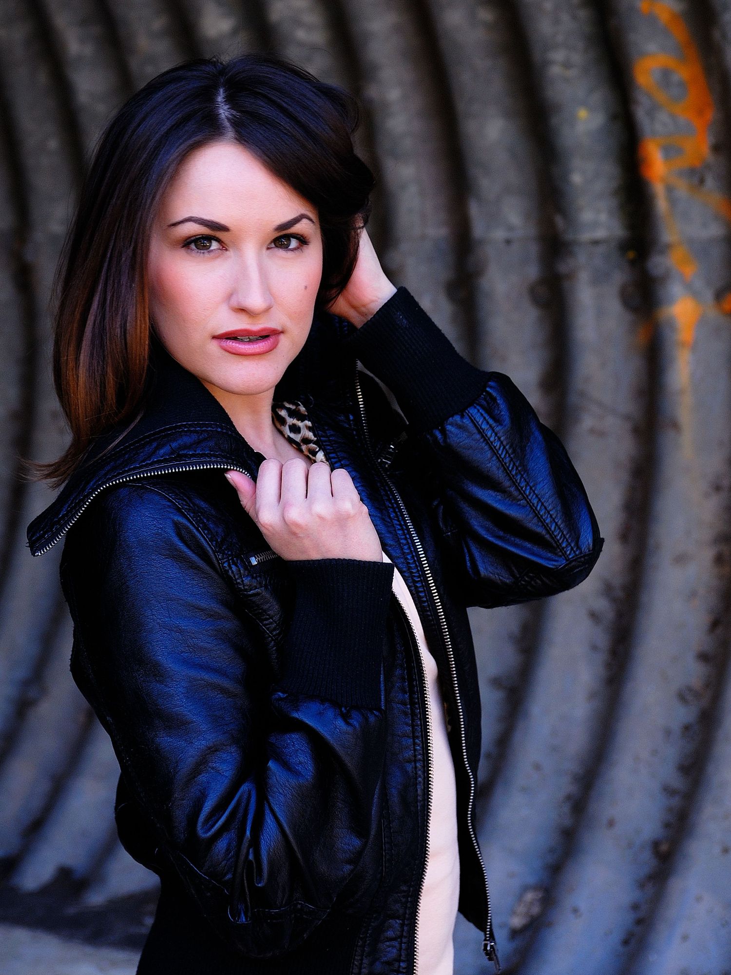 A woman in a black leather jacket poses confidently against a corrugated metal wall backdrop.