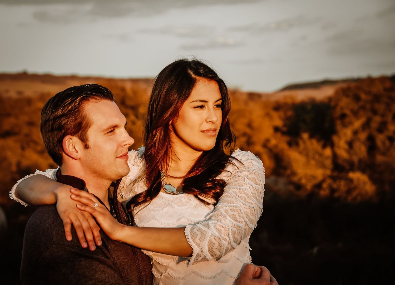 Couple embracing outdoors at golden hour, woman hugging man from behind in warm autumn landscape.
