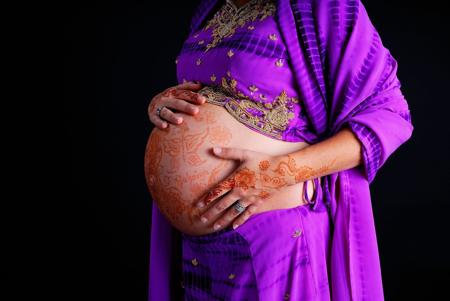 Pregnant woman in purple sari with henna mehndi on hands cradling bare baby bump against dark background.