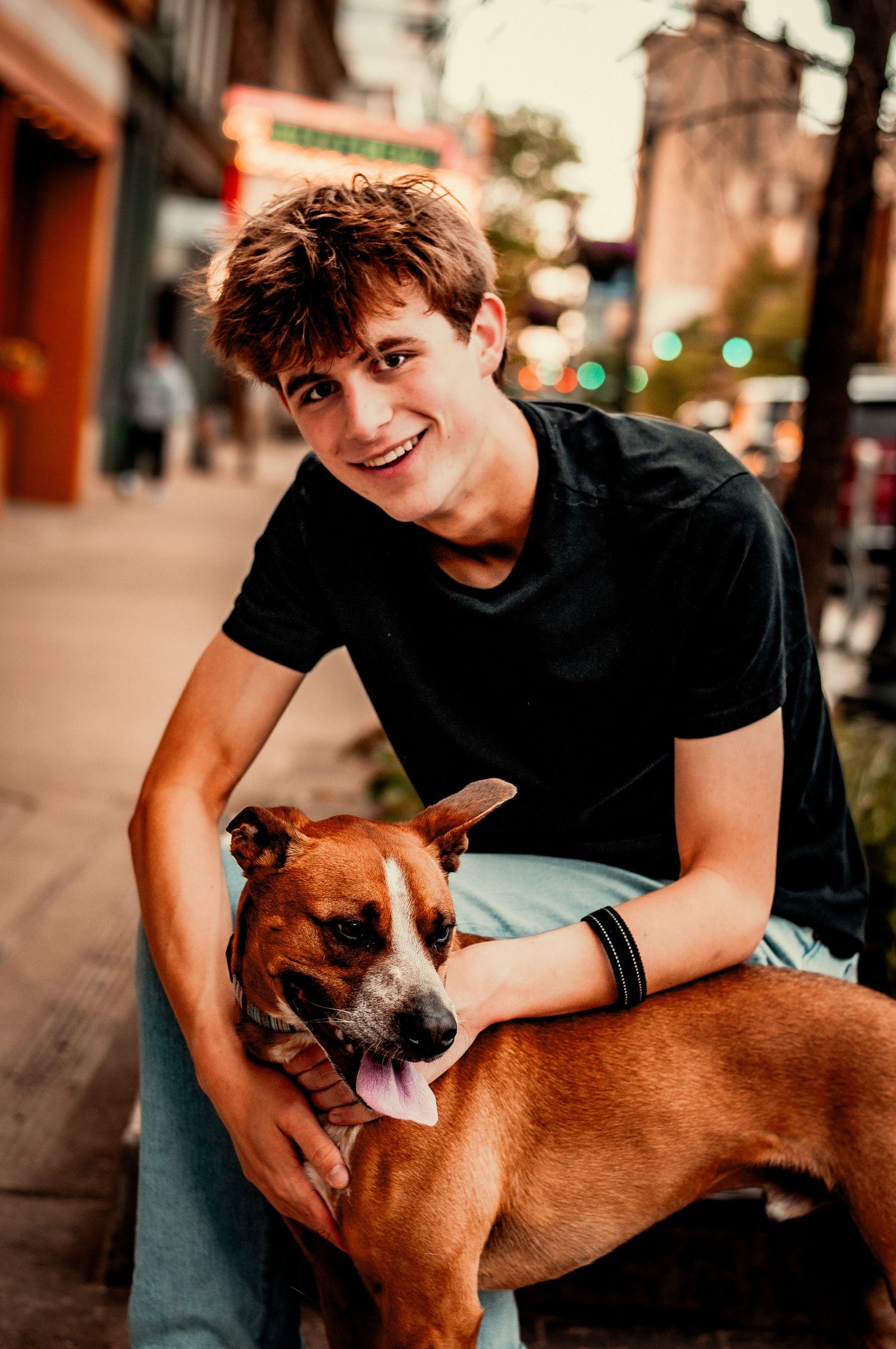 Smiling young man in black t-shirt sitting outdoors on urban sidewalk, petting a happy brown boxer mix dog.