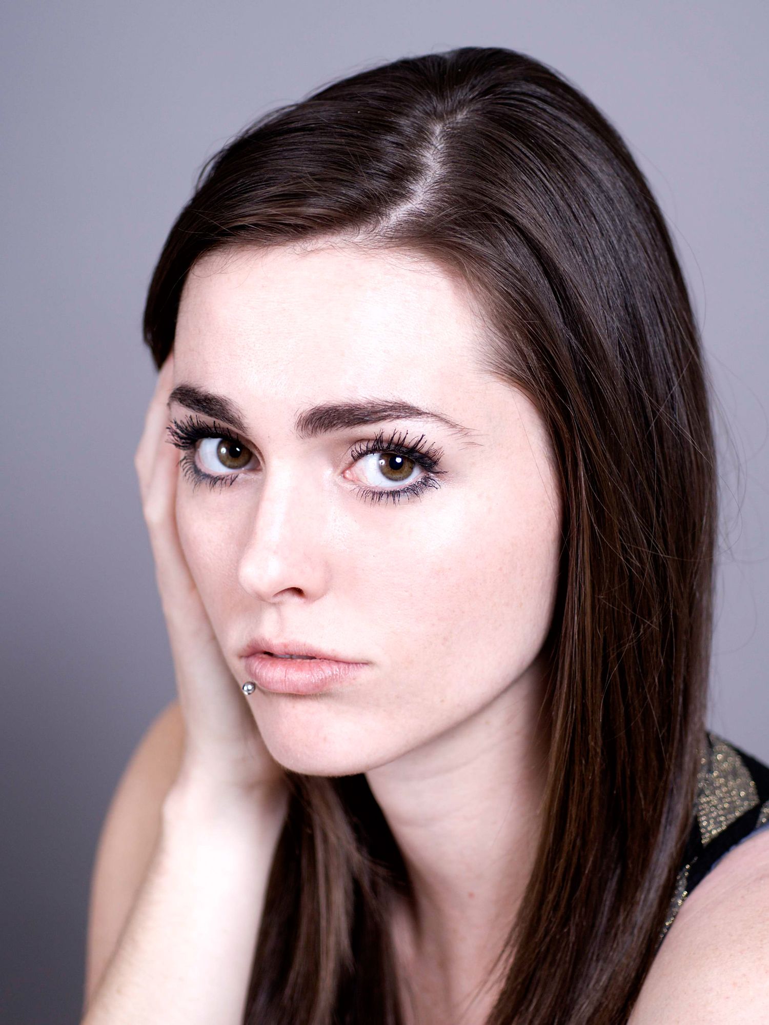 Young woman with dark brown hair and striking eyes poses thoughtfully with hand near face against gray background.