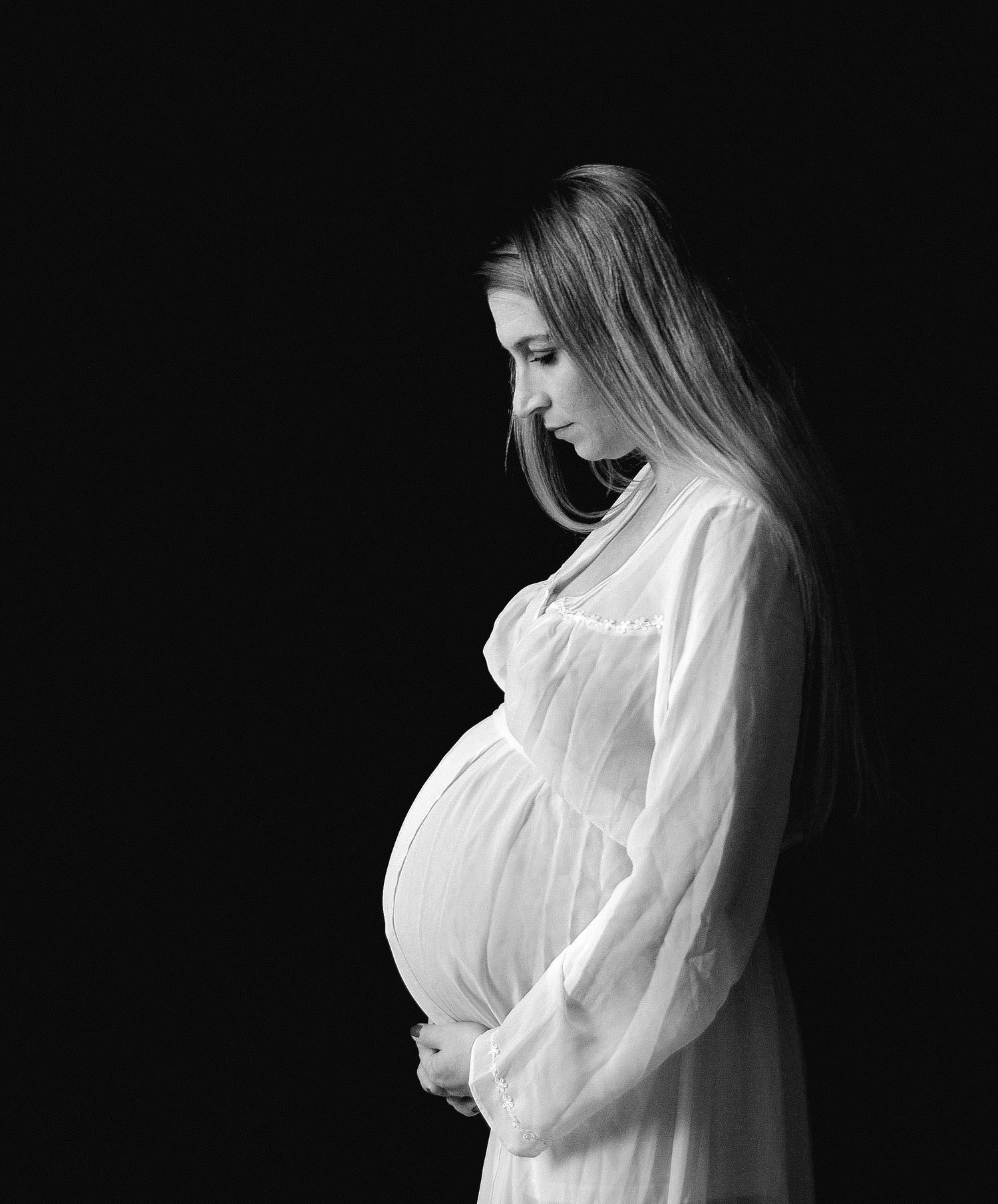 Black and white maternity photo of a pregnant woman in white dress cradling her baby bump against dark background.