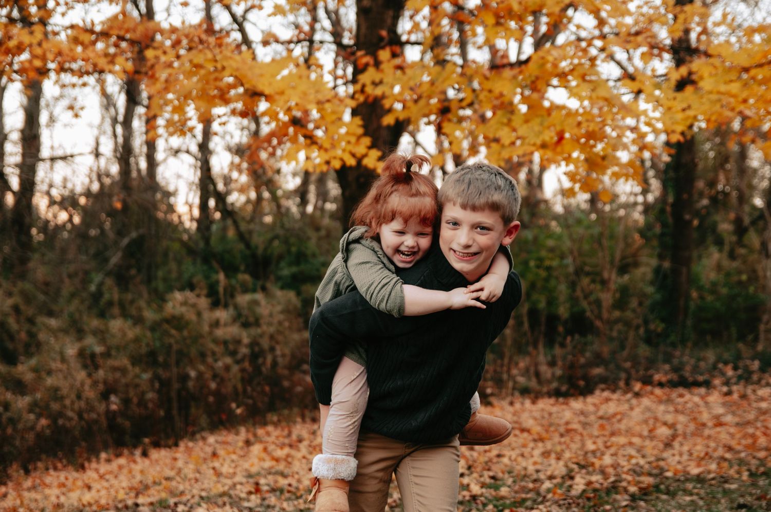 Two children enjoy a piggyback ride outdoors among vibrant autumn foliage in a warm fall forest setting.