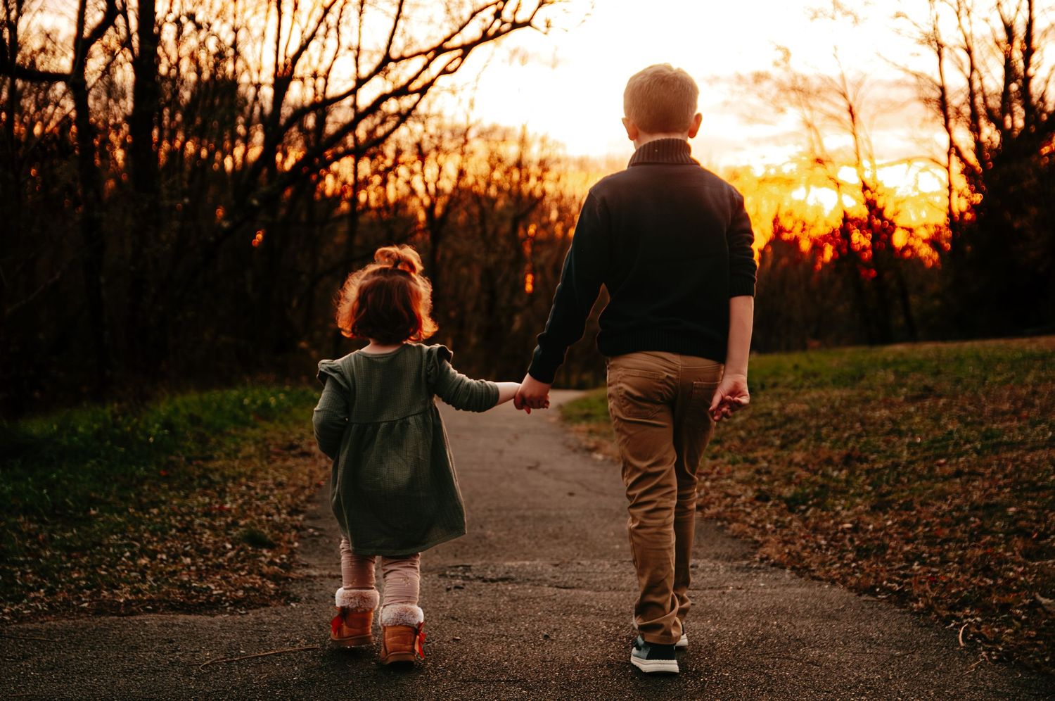 Two children holding hands walk along a path at sunset surrounded by autumn trees.