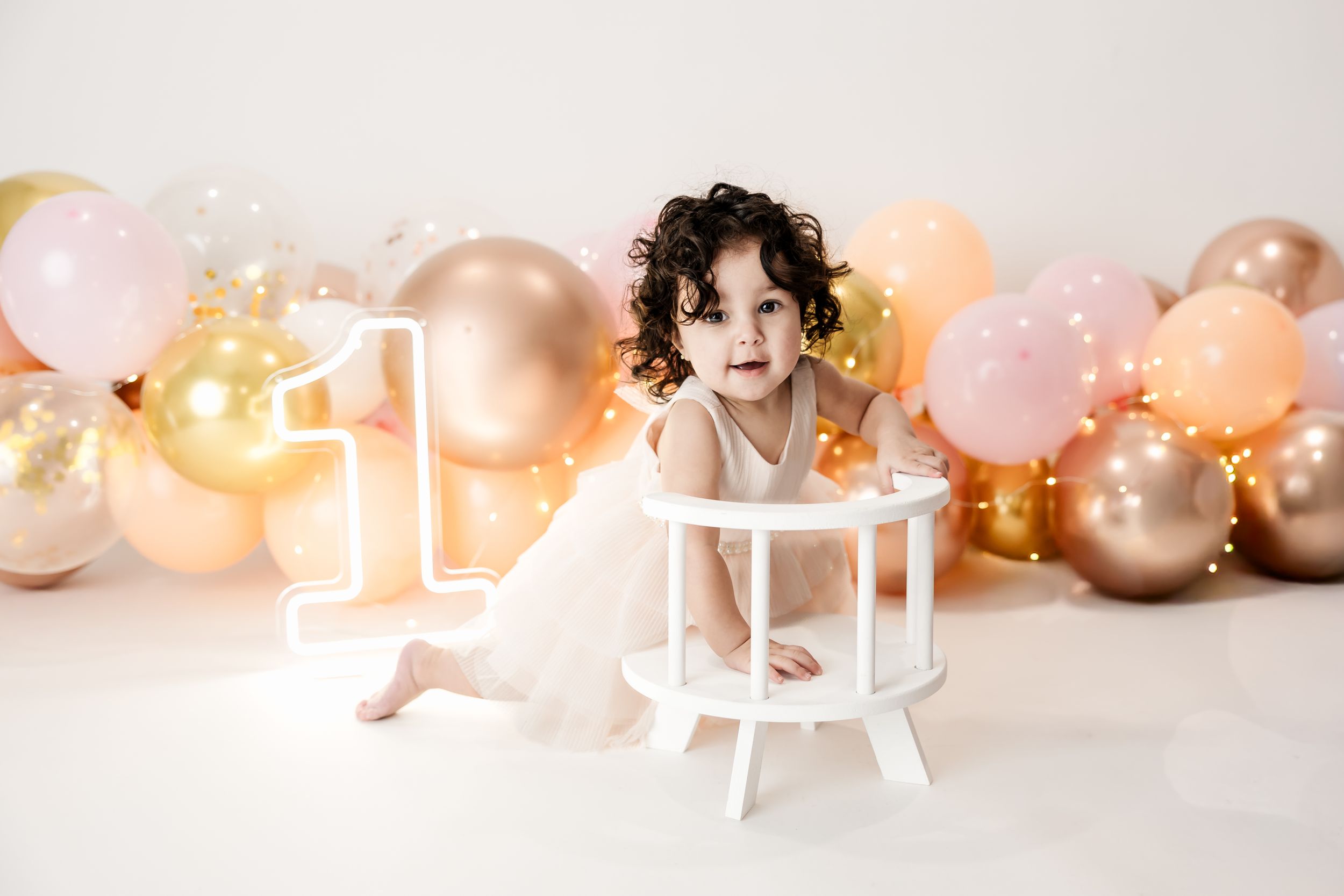 Cute toddler girl in white tutu dress sitting by number one sign at first birthday balloon photo shoot.