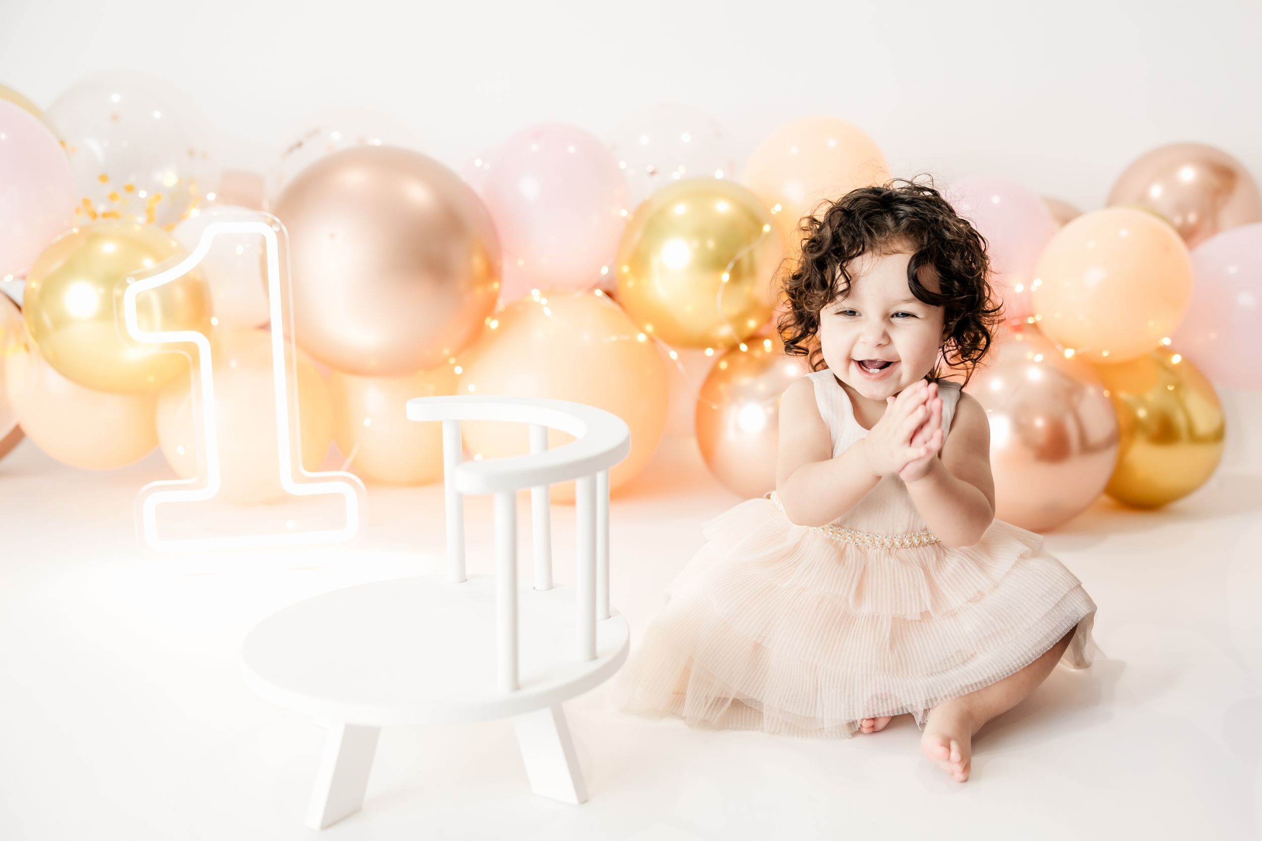 Happy toddler girl in white dress clapping hands joyfully surrounded by gold and pink balloons at birthday.