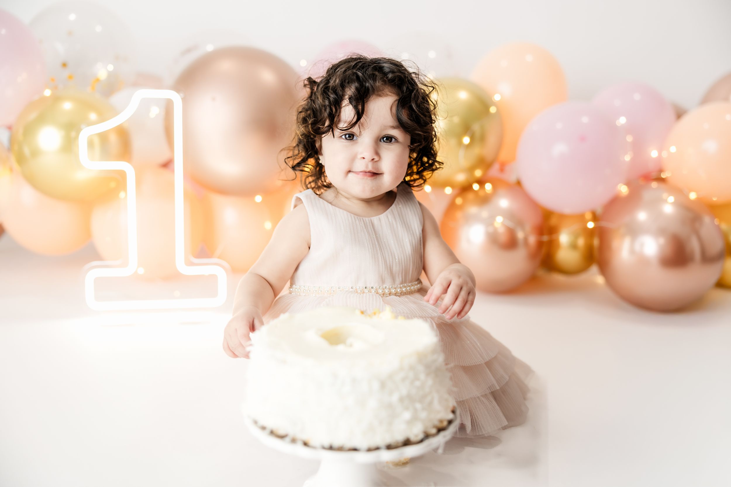Curly-haired toddler girl in white dress touching first birthday smash cake with gold balloon backdrop.
