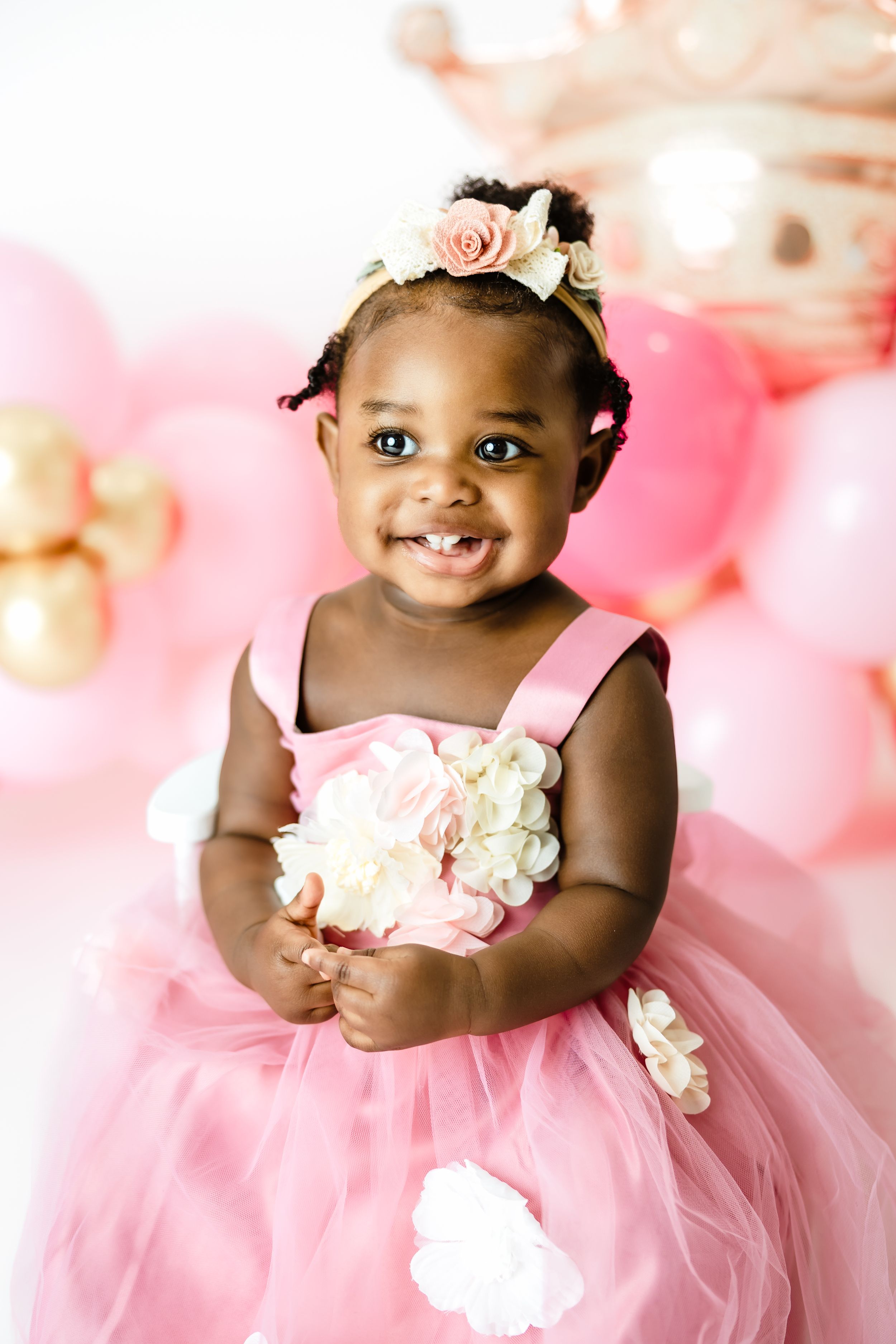 A joyful baby girl in a pink tutu dress with floral headband smiles among pink and gold balloons.