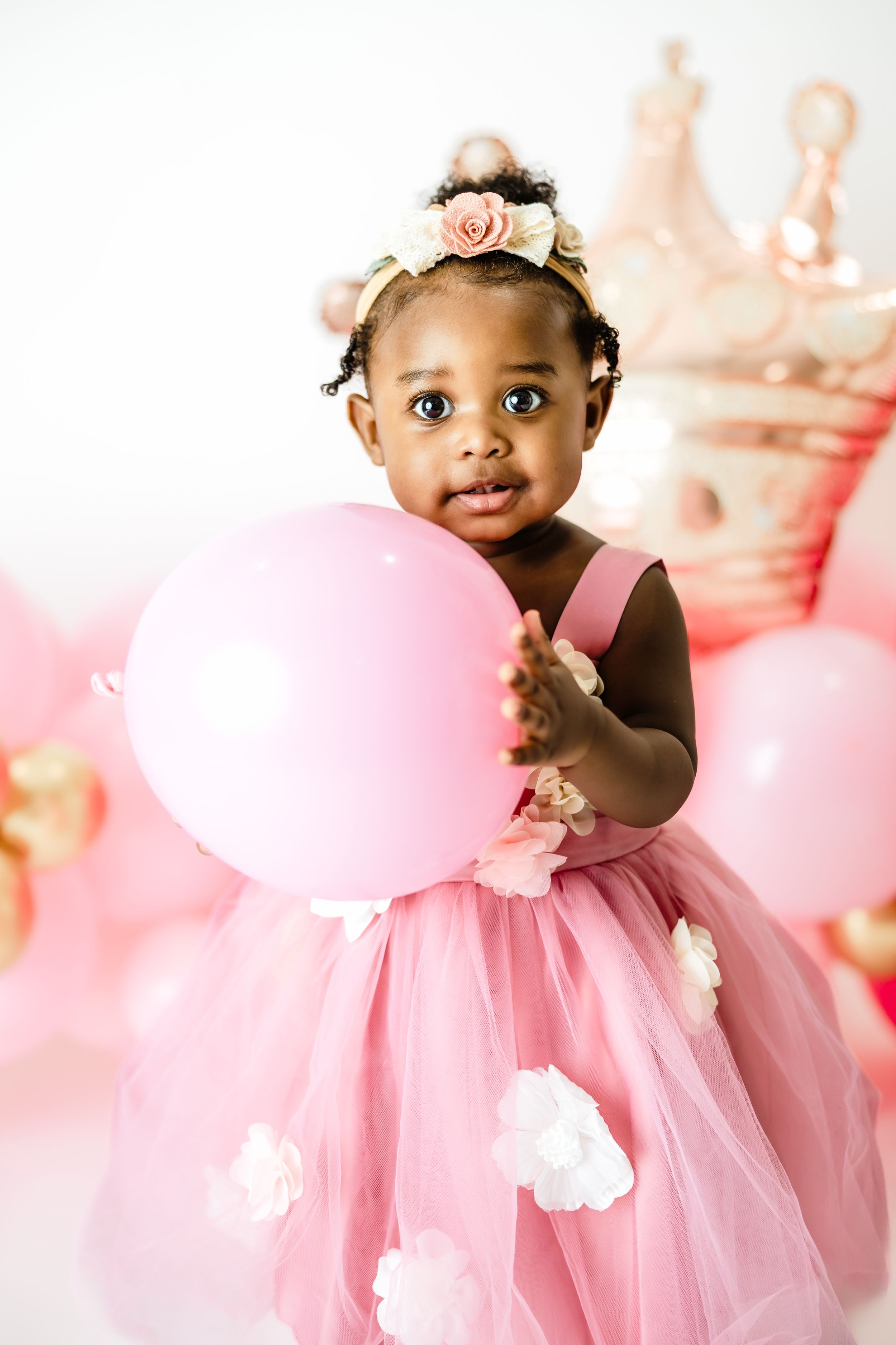 A cute baby girl in a pink tulle dress holds a pink balloon surrounded by birthday party decorations.