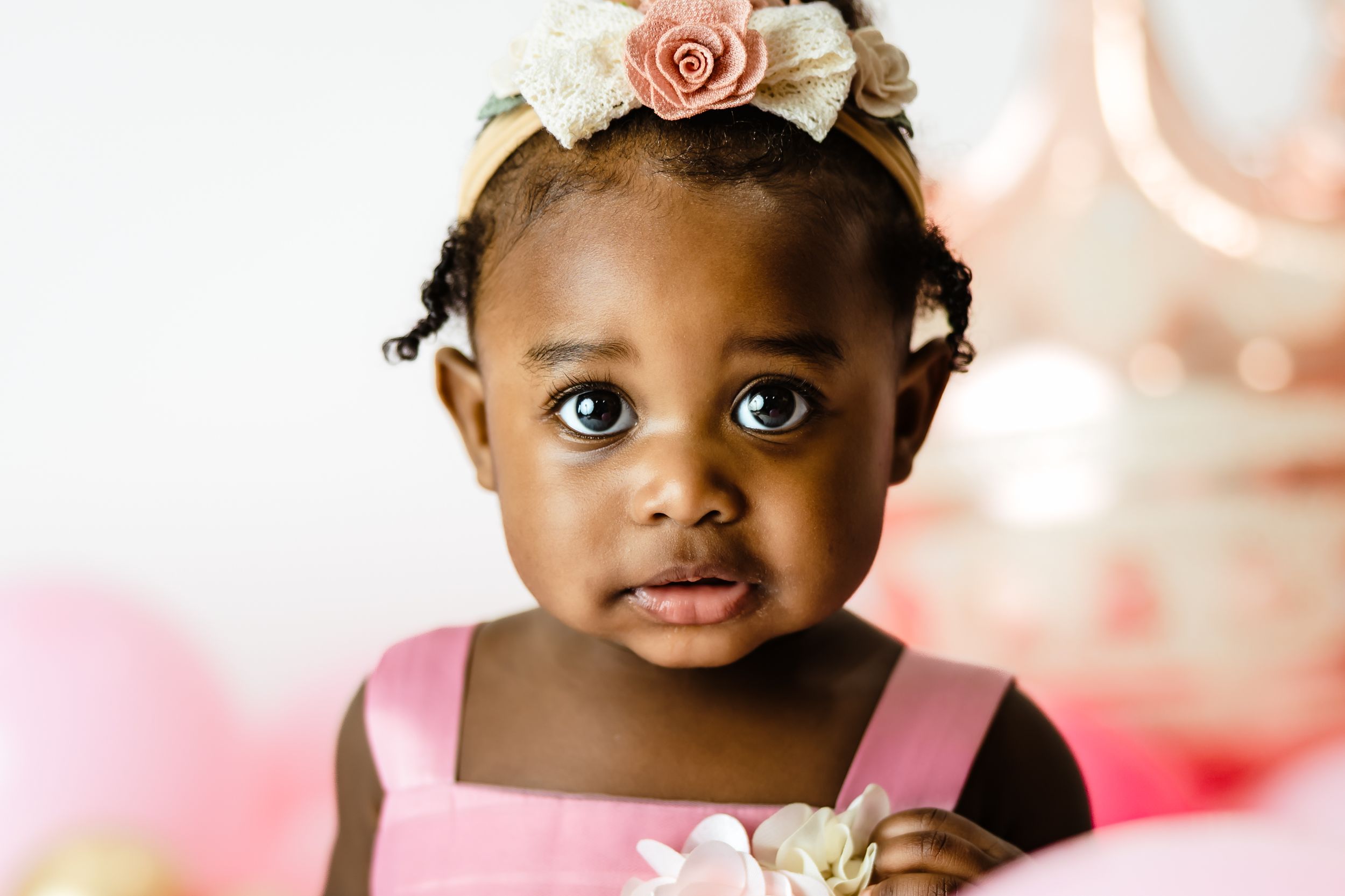 A close-up portrait of an adorable baby girl wearing a floral headband and pink dress.