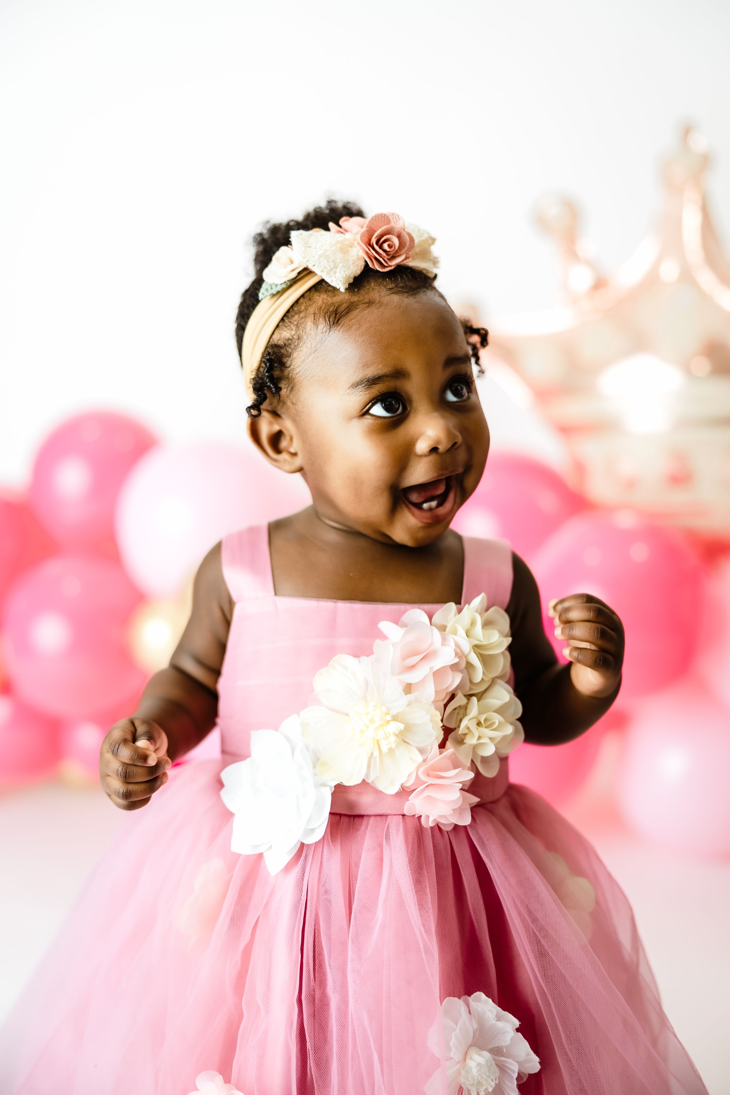 Adorable Black baby girl in pink tutu dress with floral headband smiling at first birthday party with pink balloons.