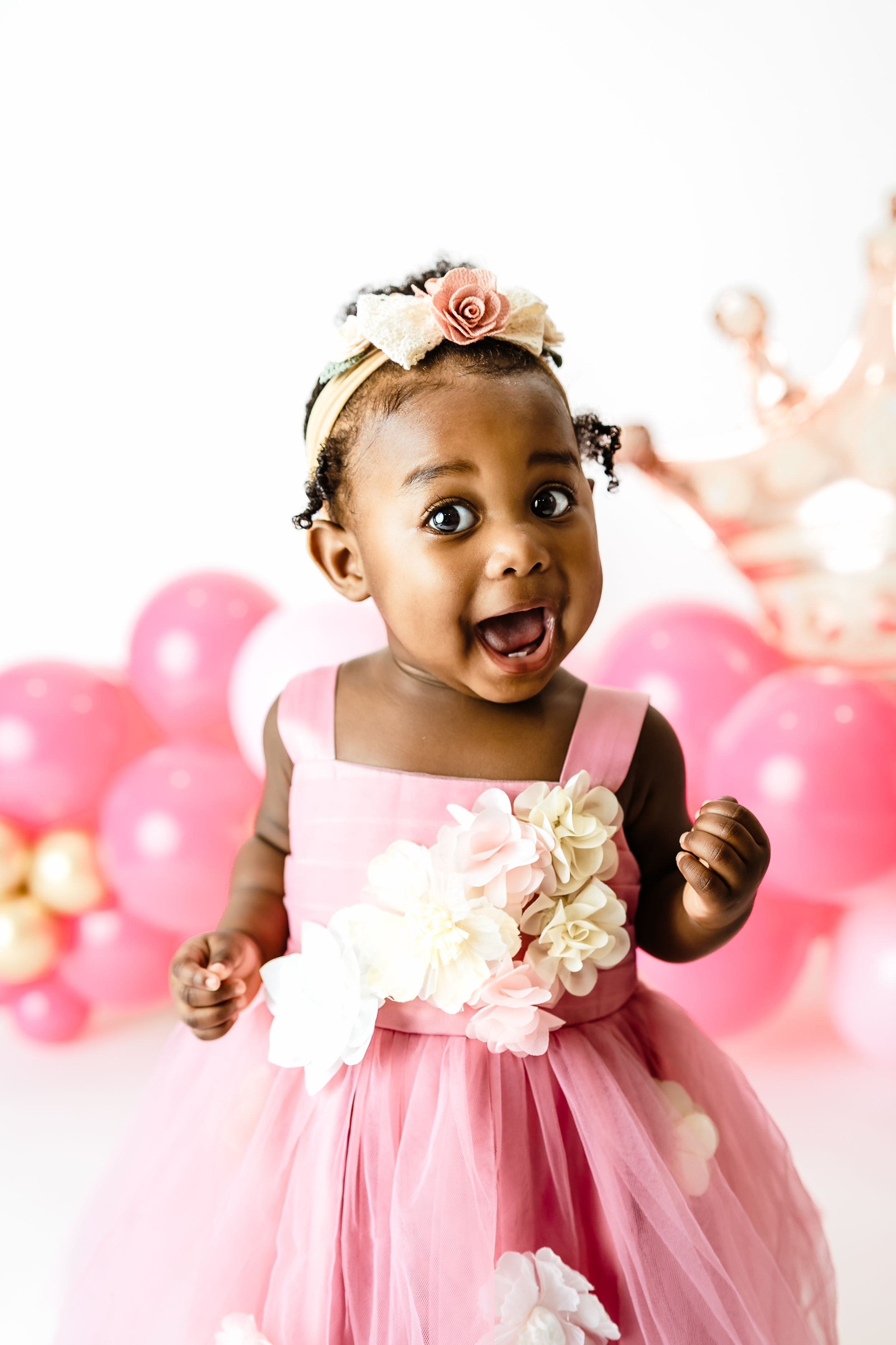 Excited Black baby girl in pink floral dress with wide eyes and open mouth at first birthday celebration.