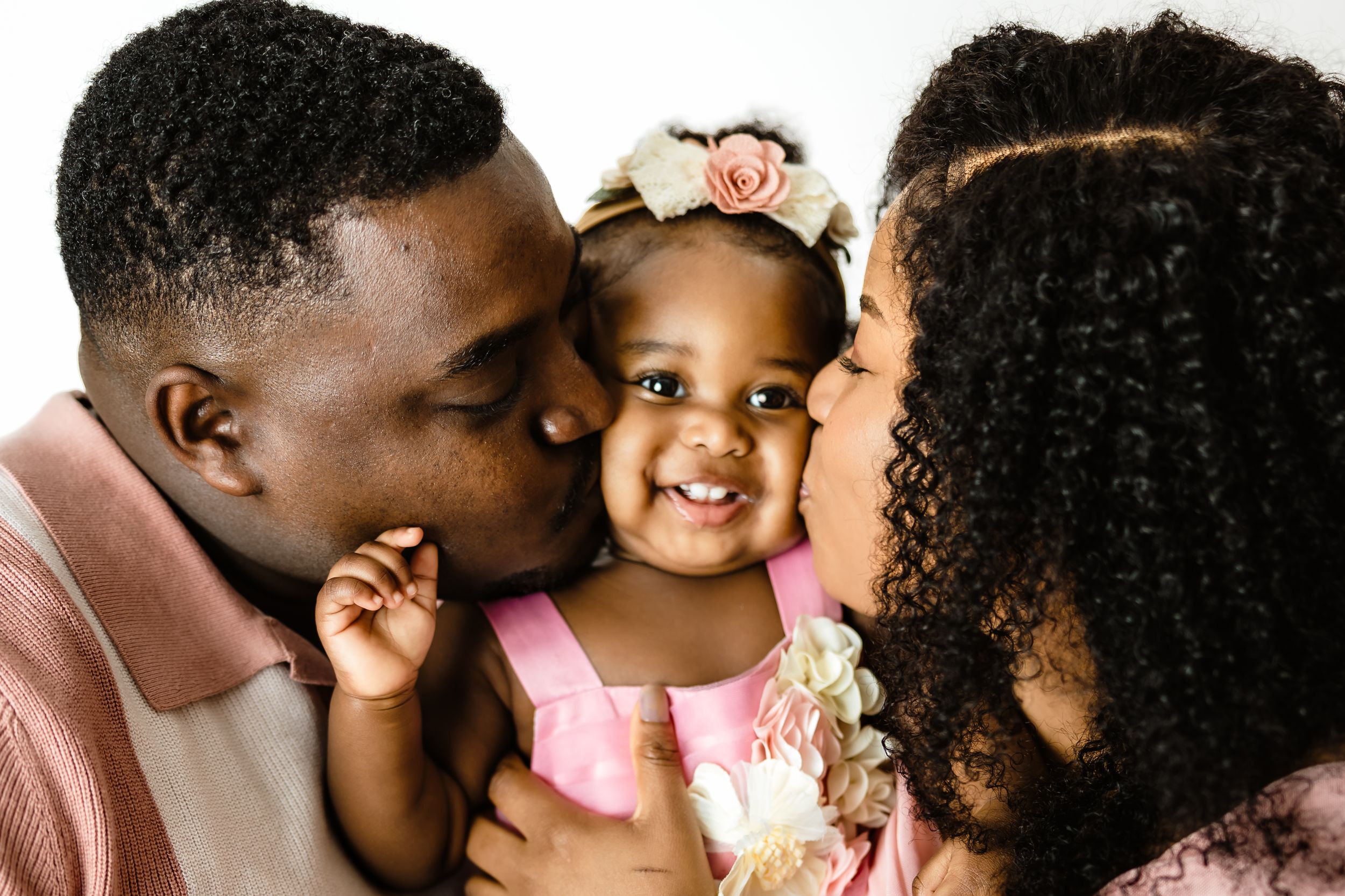 Loving Black parents kissing their smiling baby girl on cheeks during first birthday photo session.
