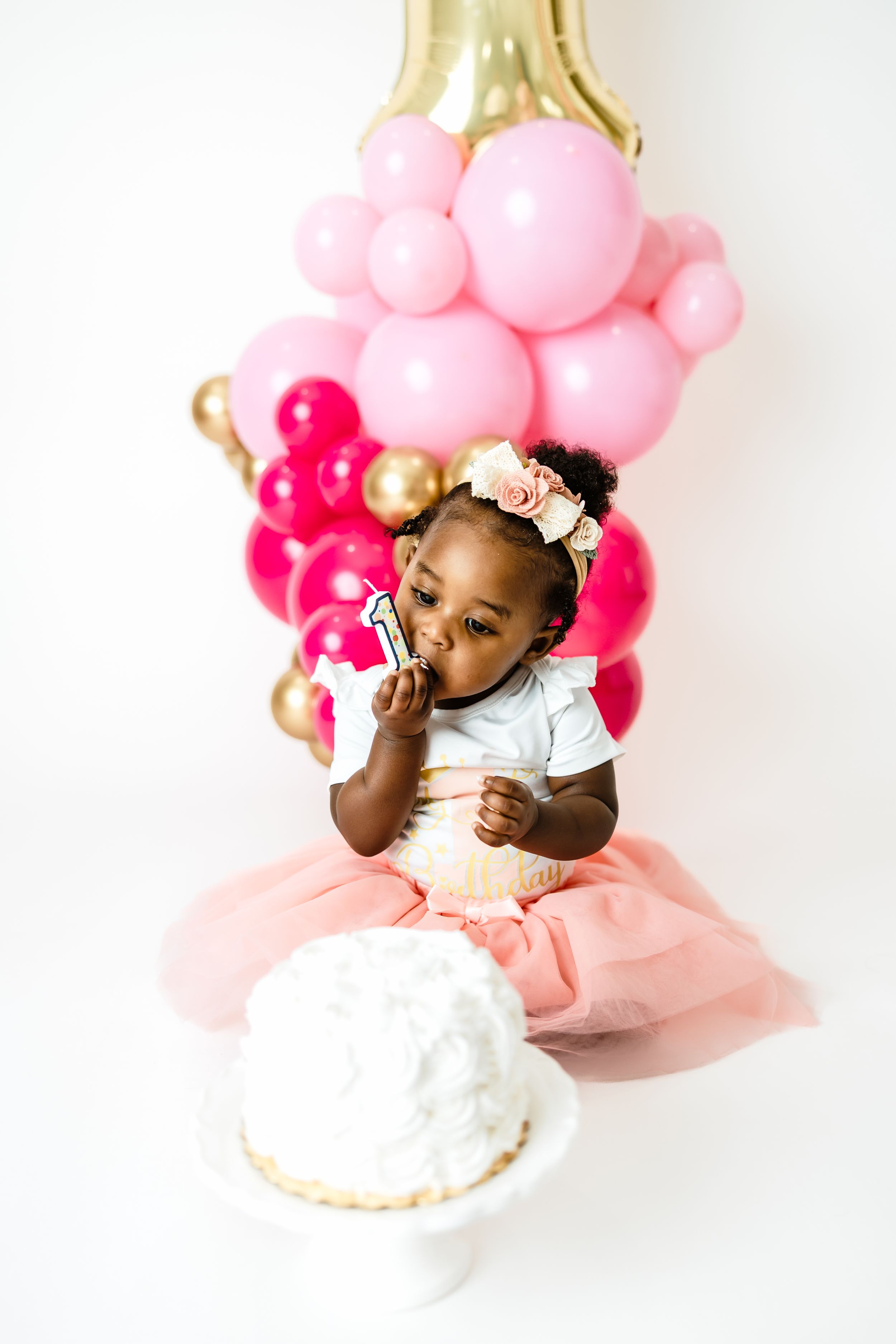 Black baby girl in pink tutu holding candle beside white smash cake at first birthday balloon backdrop.
