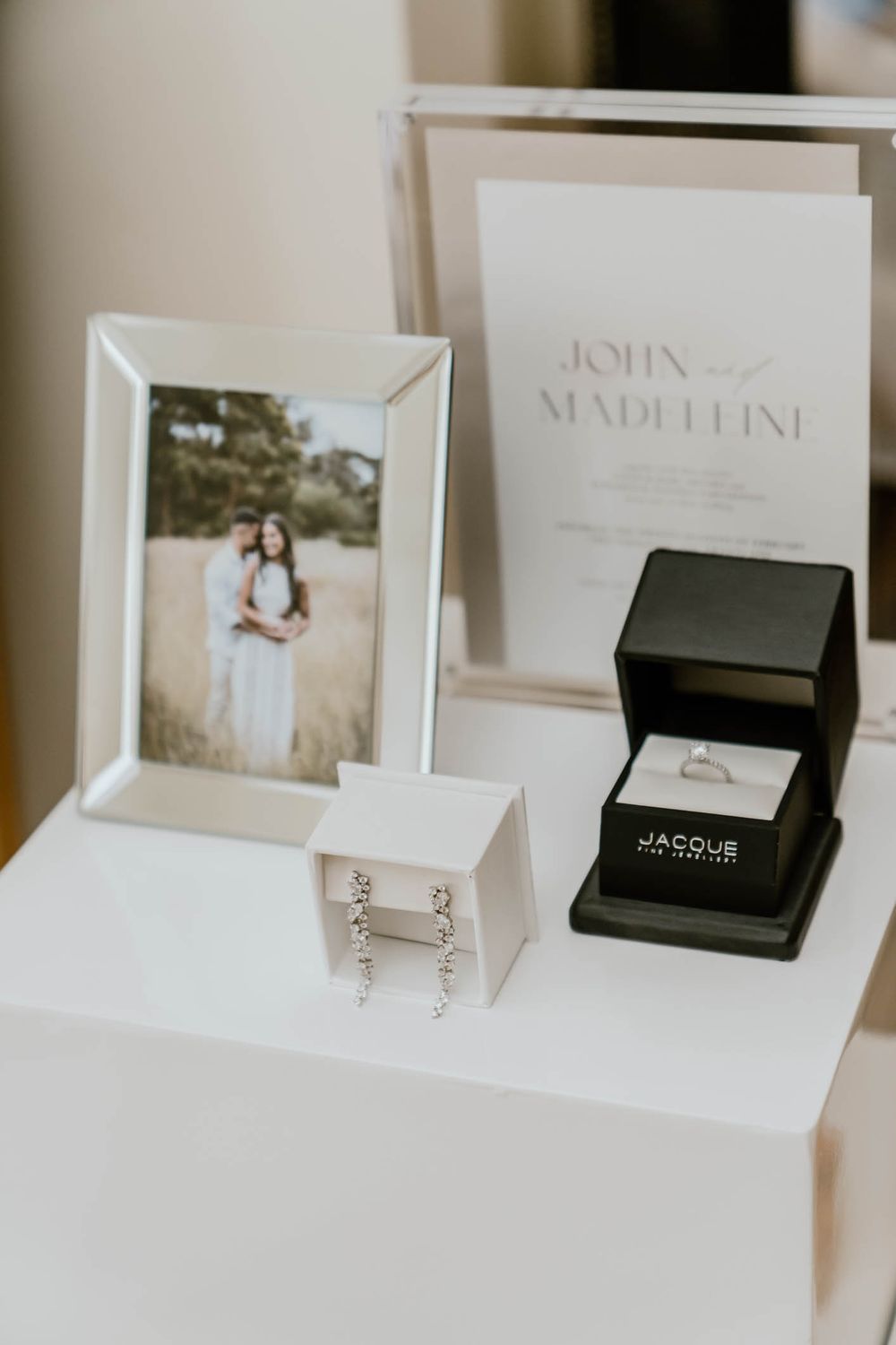 Wedding flat lay featuring an engagement photo, diamond ring in Jacobs box, earrings, and John and Madeleine invitation.