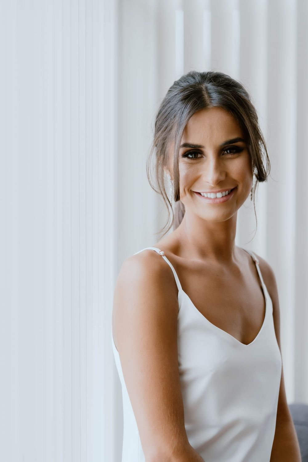 Smiling bride in a white satin slip dress with elegant updo hairstyle and natural makeup poses near a window.