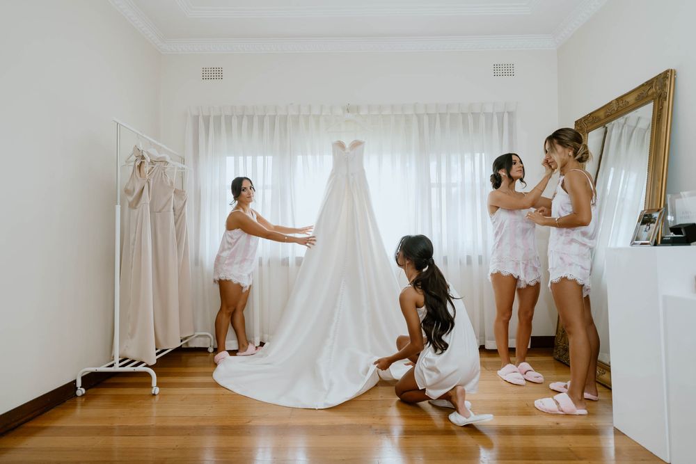 Bridesmaids in white robes help arrange a elegant wedding gown on a rack in a bright, airy bridal suite.