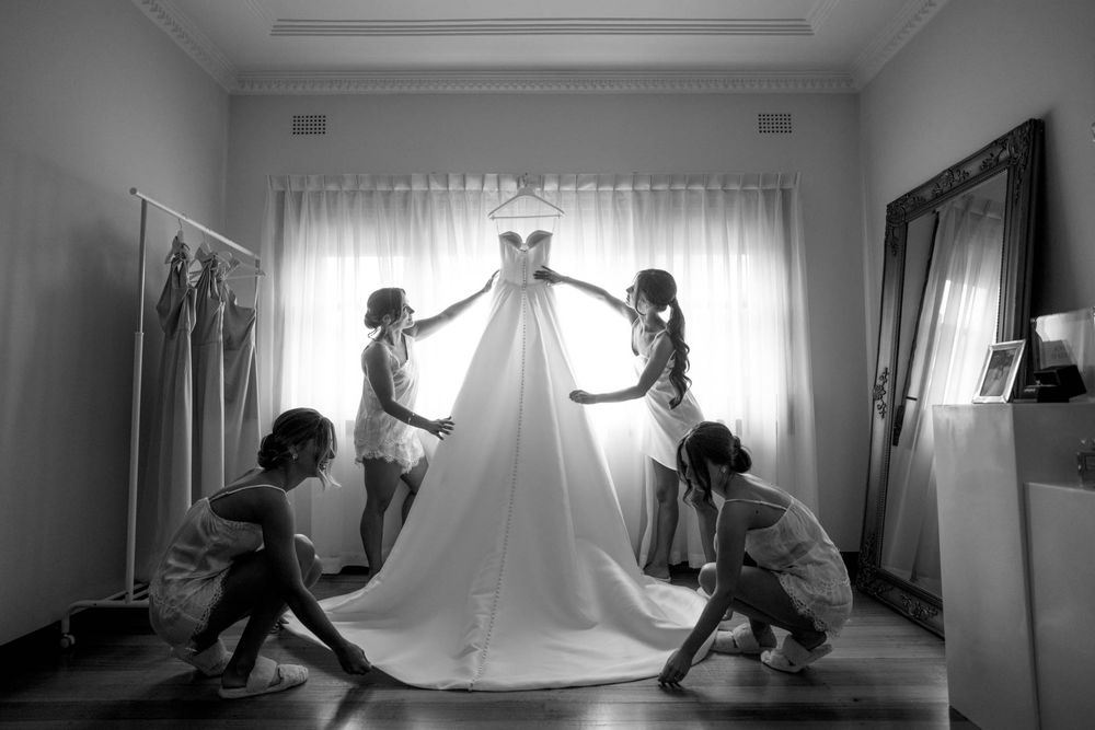 Four women arrange a stunning white wedding gown in a sunlit room in this elegant black and white bridal preparation photo.