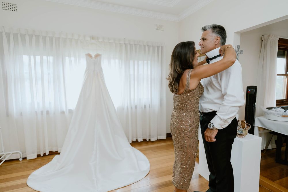 Bride and father share an emotional embrace in a bright room with a elegant white wedding gown displayed in the background.
