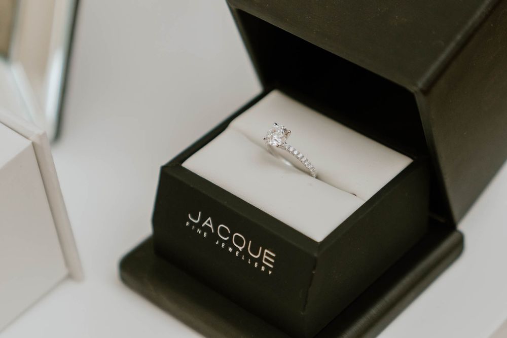 Diamond solitaire engagement ring displayed in a black Jacque The Jeweller ring box on a white surface.