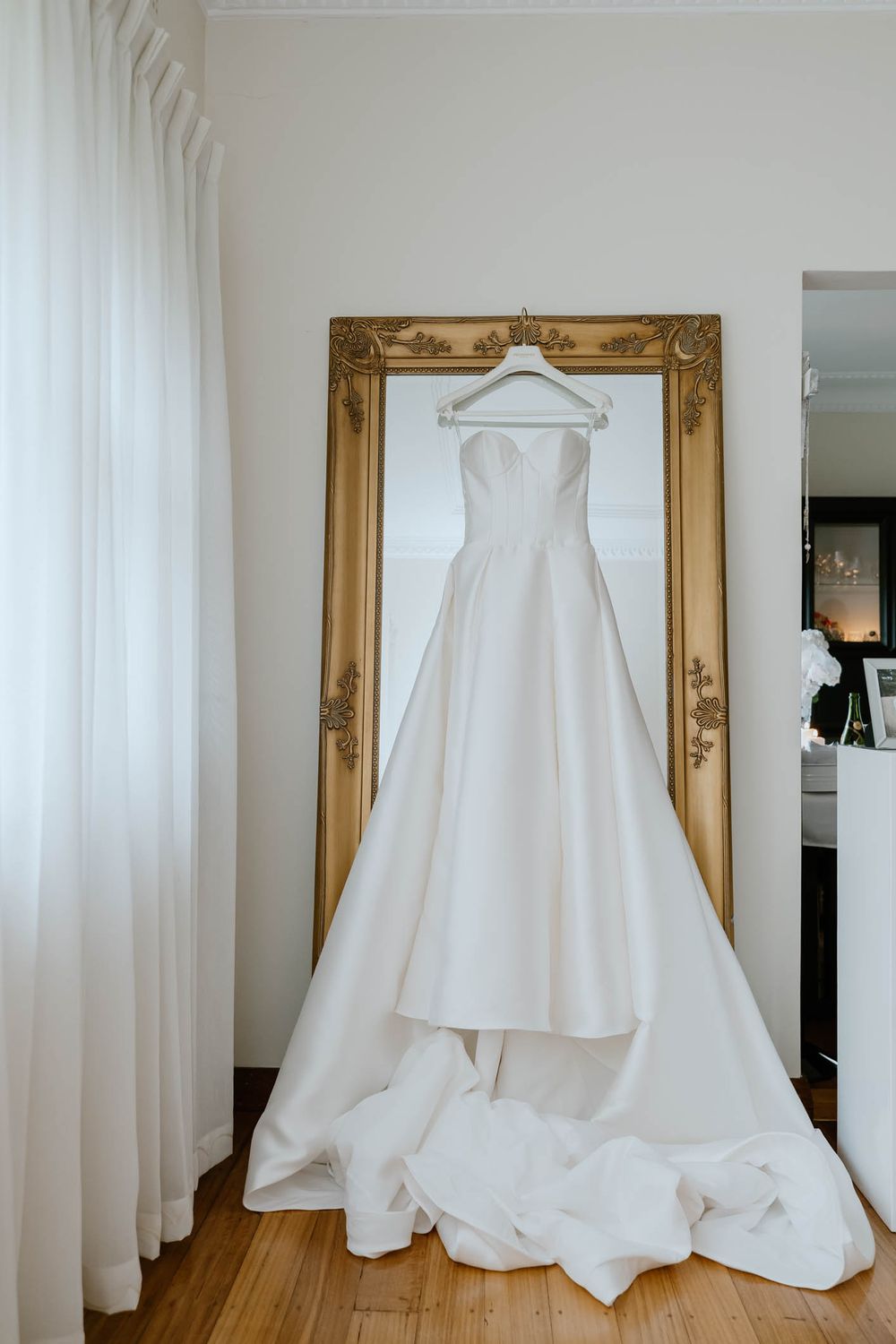 Elegant strapless white ball gown wedding dress with full skirt hanging on a gold ornate mirror frame.