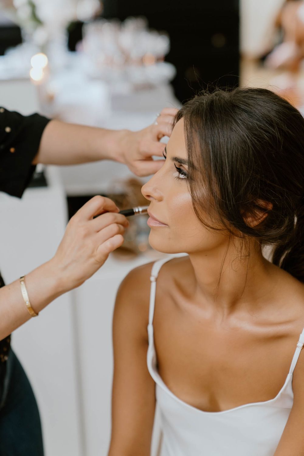 Makeup artist applying finishing touches to a brunette bride in a white dress before her wedding.