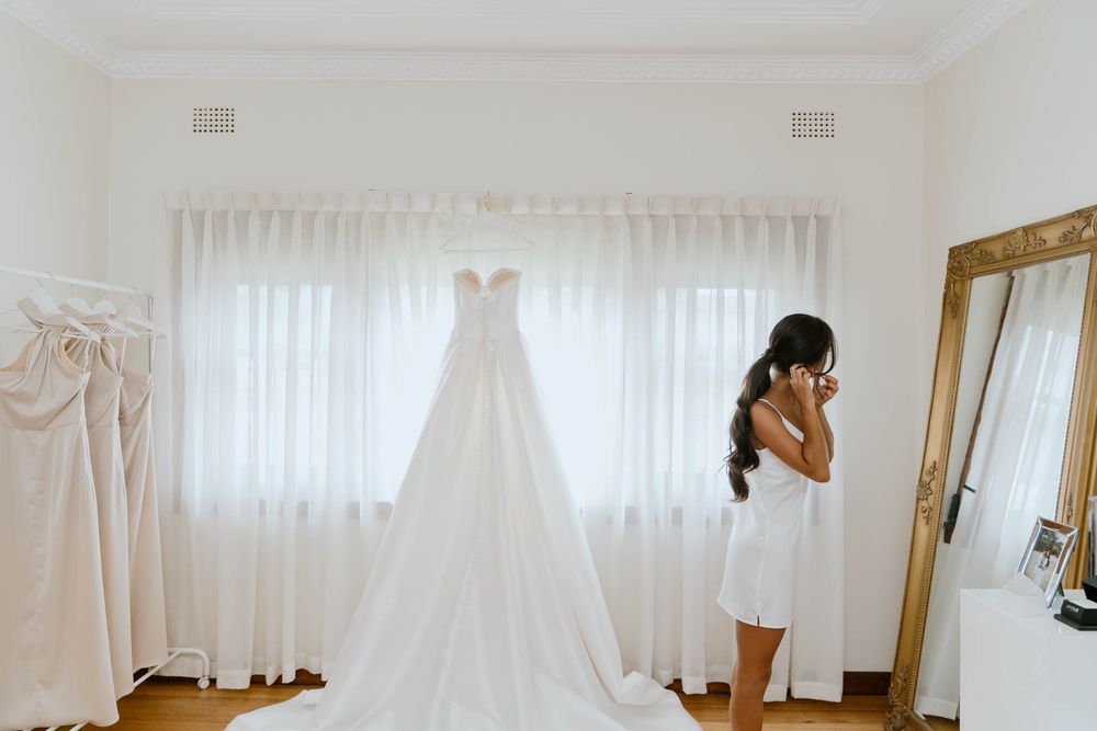 Bride in white robe admires her elegant strapless wedding gown hanging by a sunlit window in a bright bridal suite.