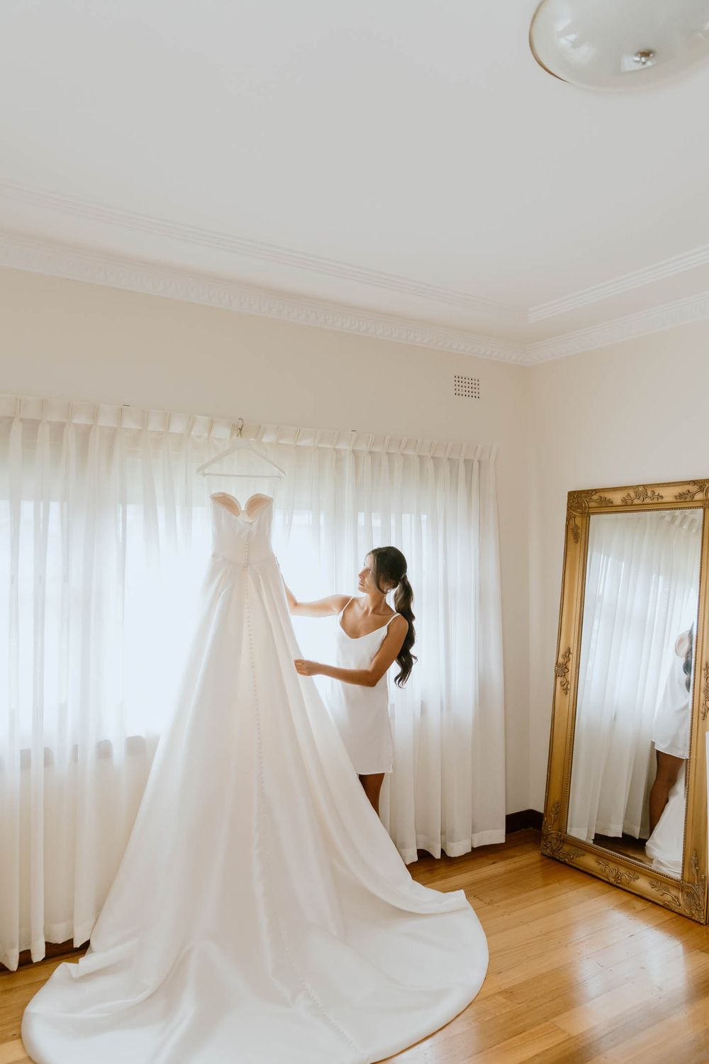 Bride in white slip dress admiring her elegant wedding gown hanging by a sunlit window with a gold mirror nearby.