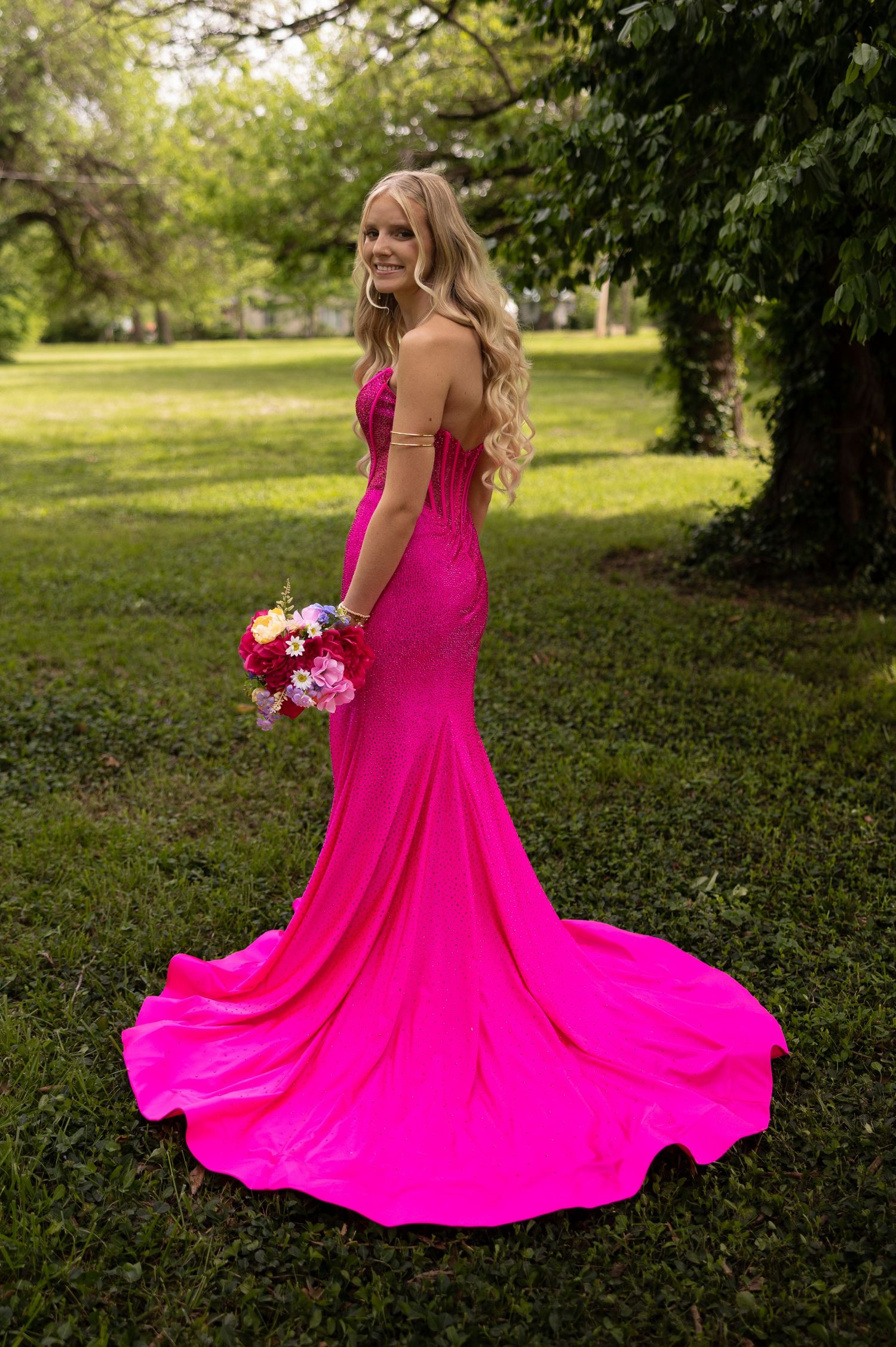 Woman in hot pink mermaid gown holding colorful bouquet, posing outdoors in lush green garden setting.