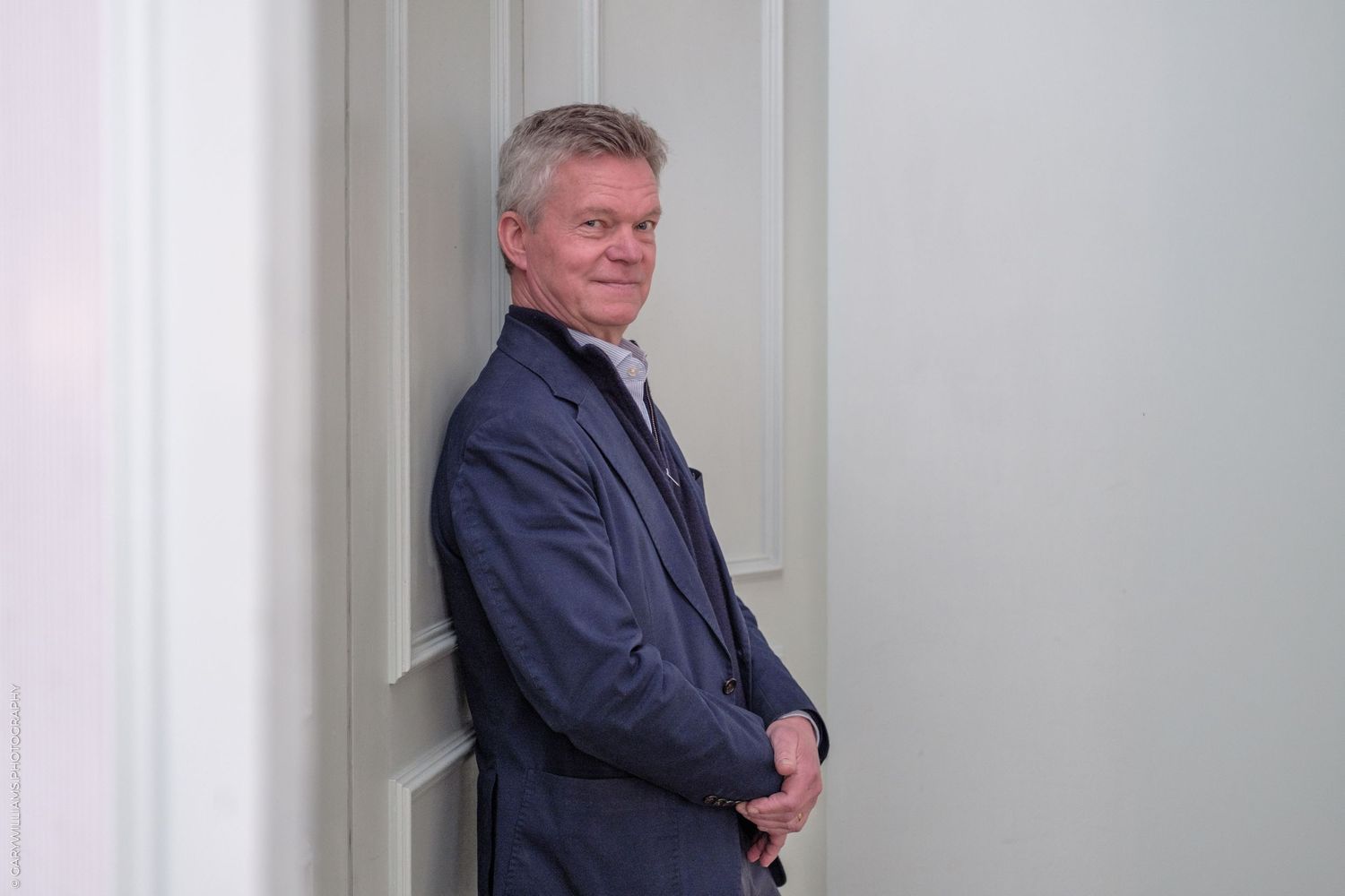 Soren Jessen of 1 Lombard Street in a blue blazer leaning against a white wall, smiling confidently indoors.