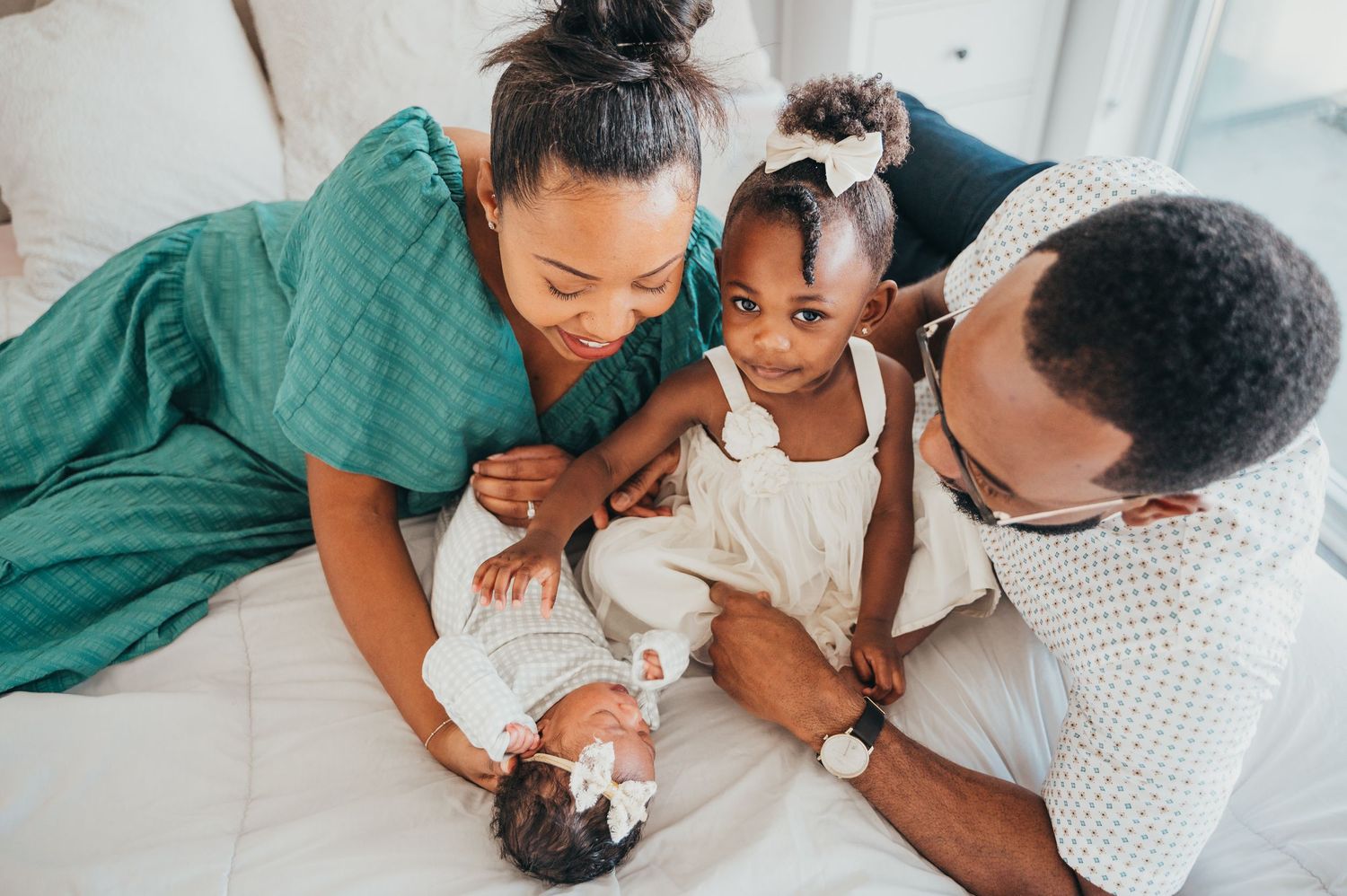 Family lays on white bed while mom and dad look and smile at newborn baby and toddler girl puts her hand on the baby and looks up