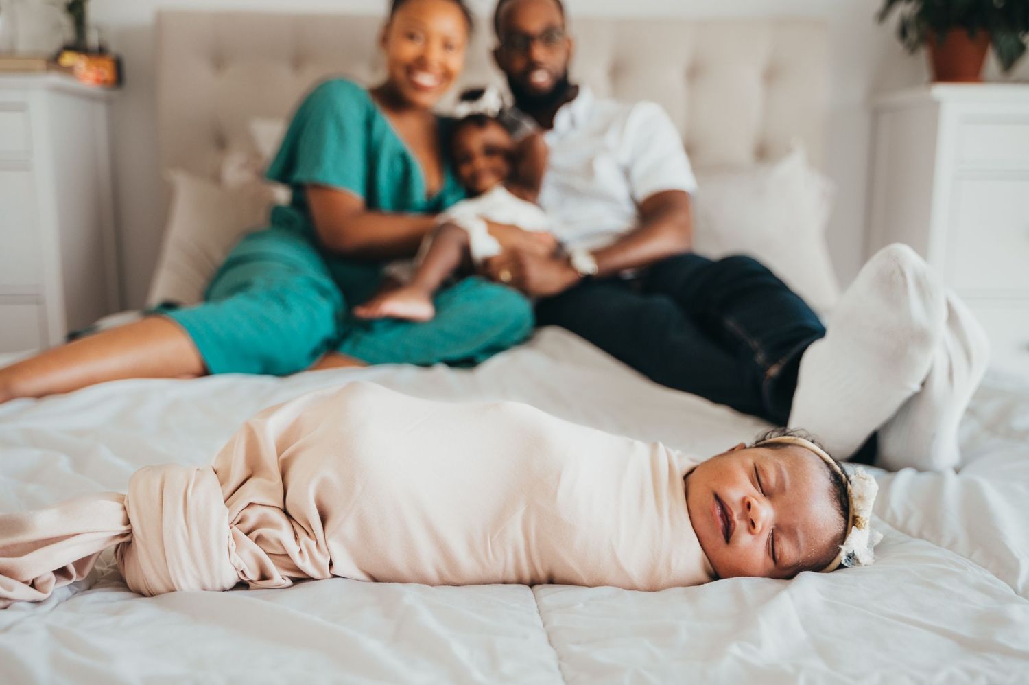 Newborn baby girls sleeping while swaddled on white bed while mom, dad and big sister look at her and smile from behind