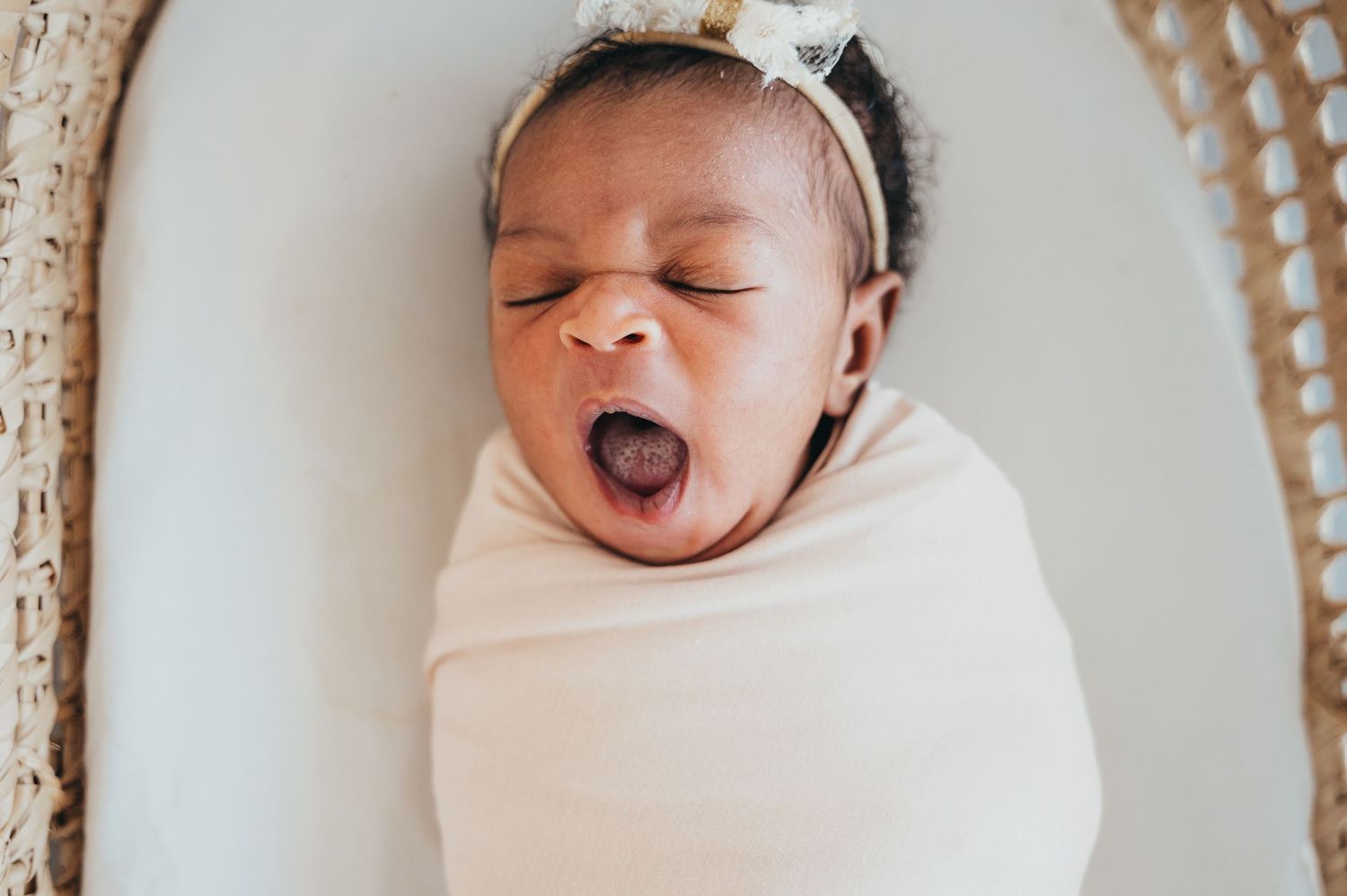 Newborn baby girl yawns while swaddled in light pink and sleeping in basket