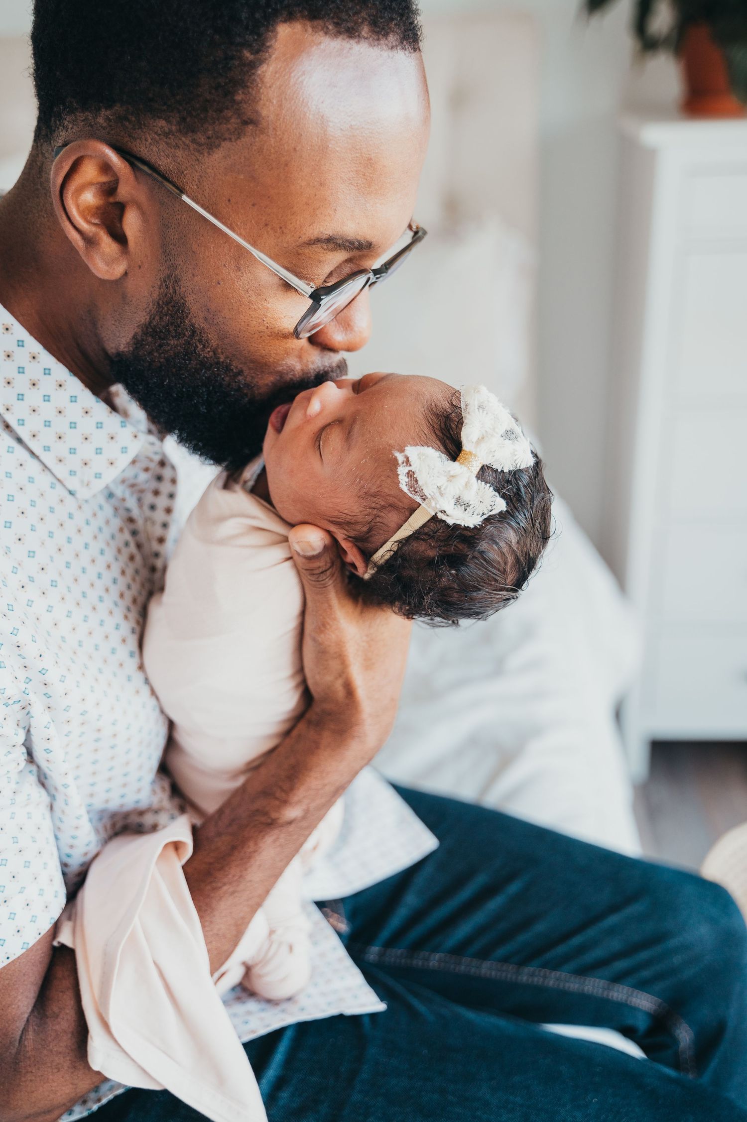 Dad holds newborn baby and kisses her head gently while sitting on a white bed