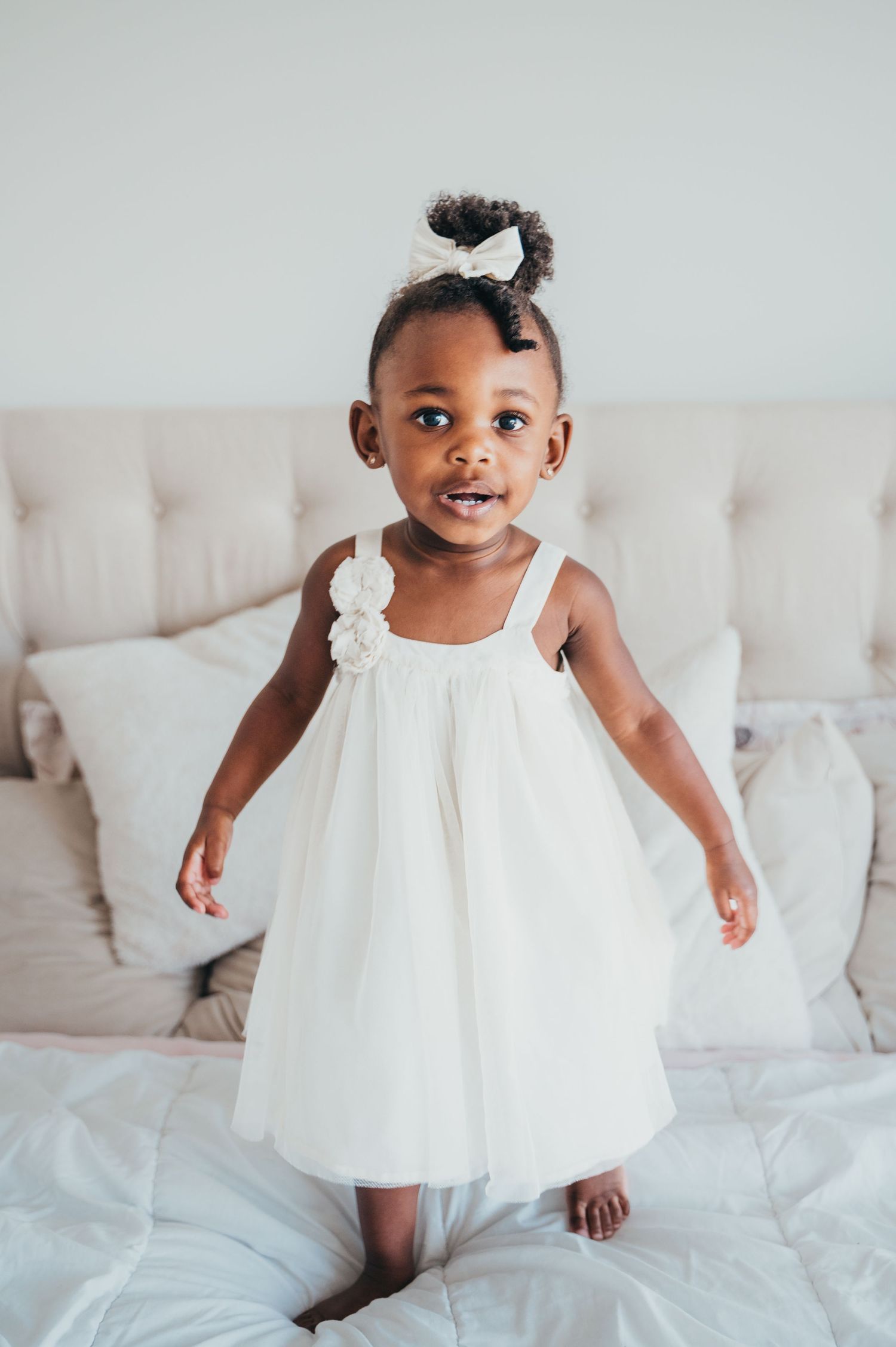 Toodler girl in white dress stands on white bed in Mississauga during newborn photo session