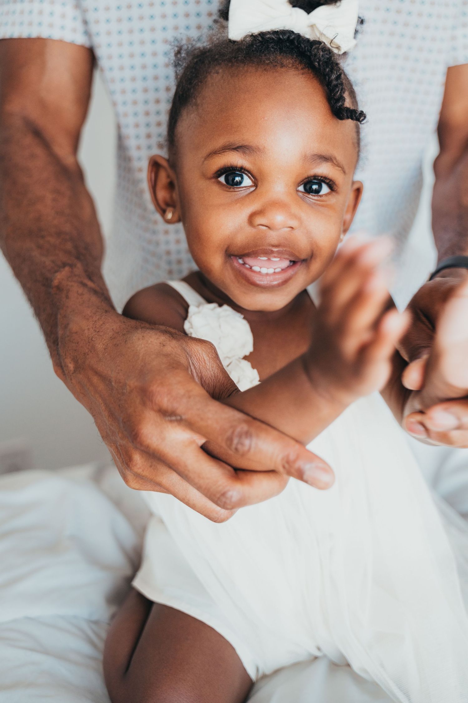 Toddler girl in white sits in front of dad while he plays with her clapping her hands on white bed in Mississauga