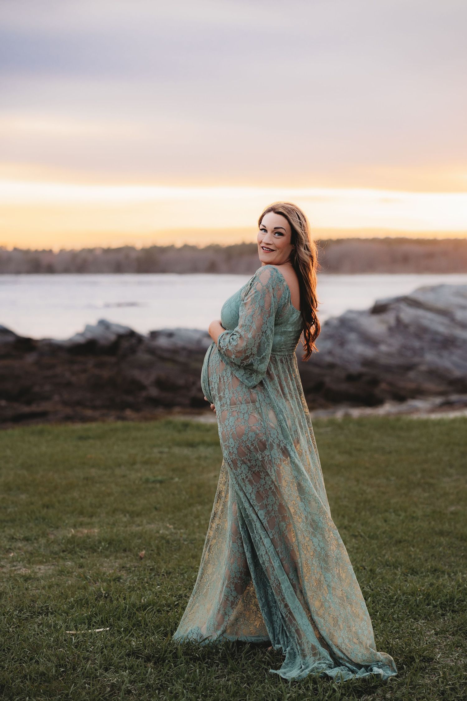 Pregnant woman in teal lace maternity gown poses outdoors at sunset near rocky shoreline, cradling her baby bump.