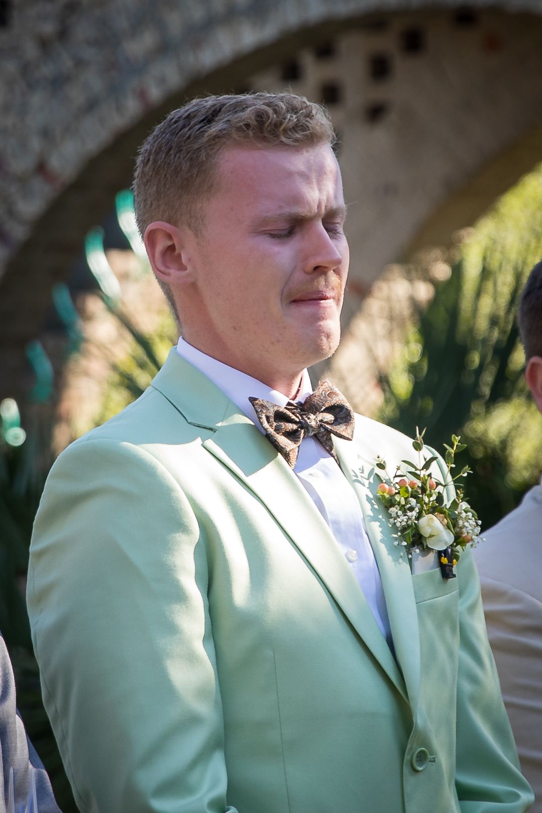 Emotional groom in mint green suit with floral boutonniere and brown bow tie, eyes closed at outdoor wedding ceremony.