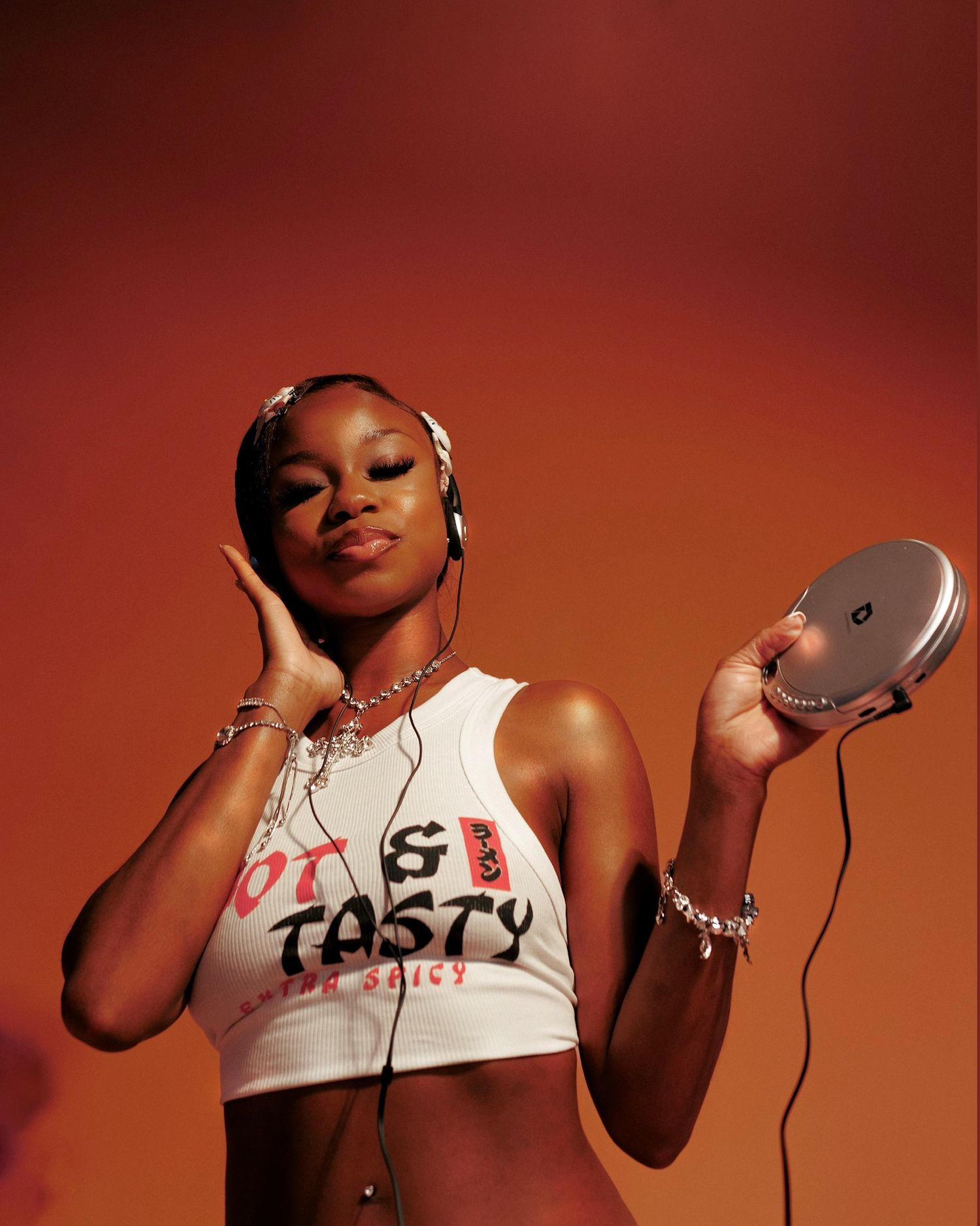 Young woman in white crop top holds portable CD player while listening to music with earphones on orange background.