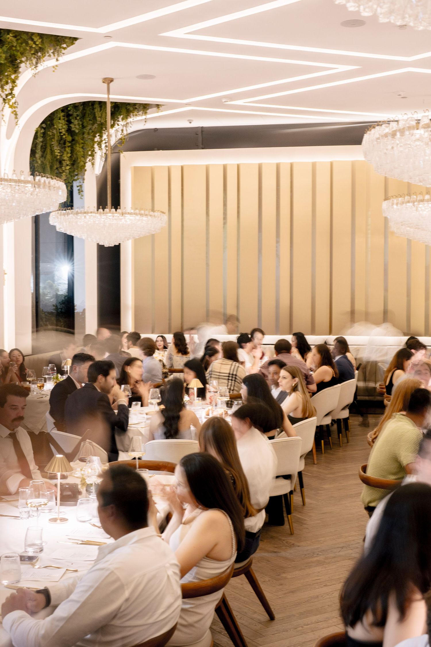 Elegant dinner party guests raising shot glasses in a toast over a white tablecloth with gold cutlery and candlelight.