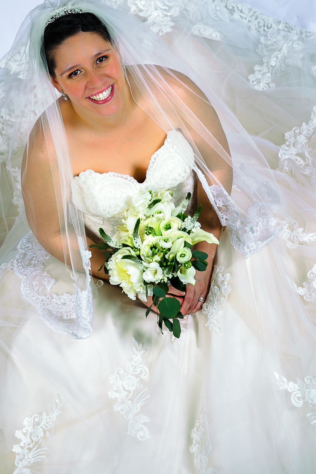 Smiling bride in white lace gown and veil holds bouquet of white flowers, looking up joyfully on her wedding day.
