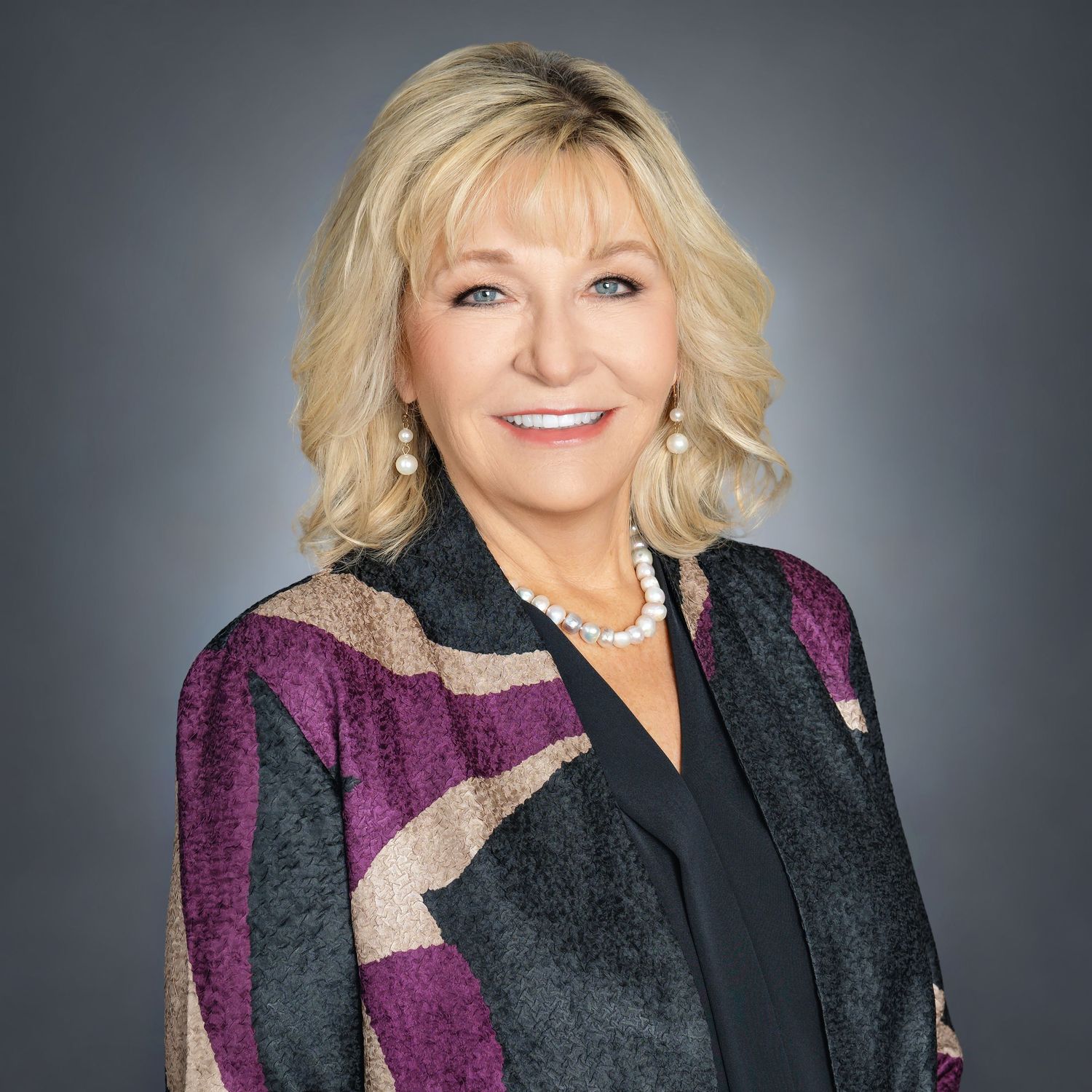 Professional headshot of a smiling blonde woman in a colorful blazer and pearl necklace against a gray background.