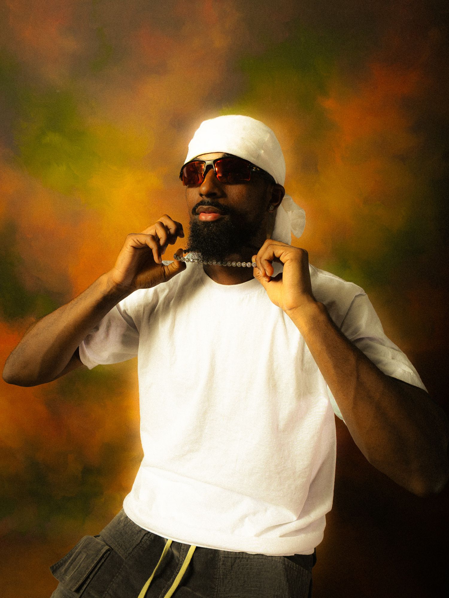 Man in white t-shirt and du-rag poses confidently wearing sunglasses and beaded necklace against colorful backdrop.