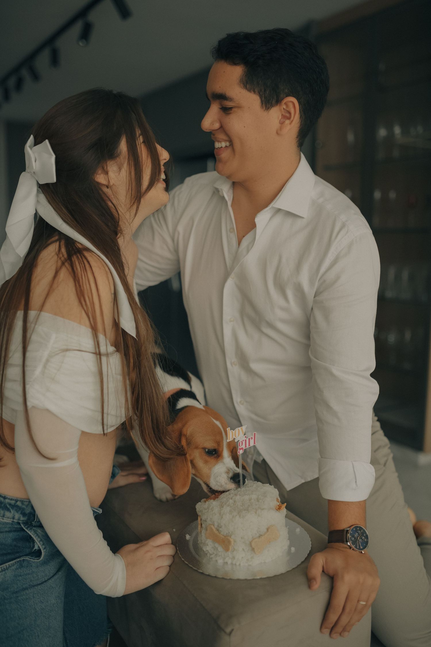 Couple celebrates with dog and cake with gender reveal decorations indoors.