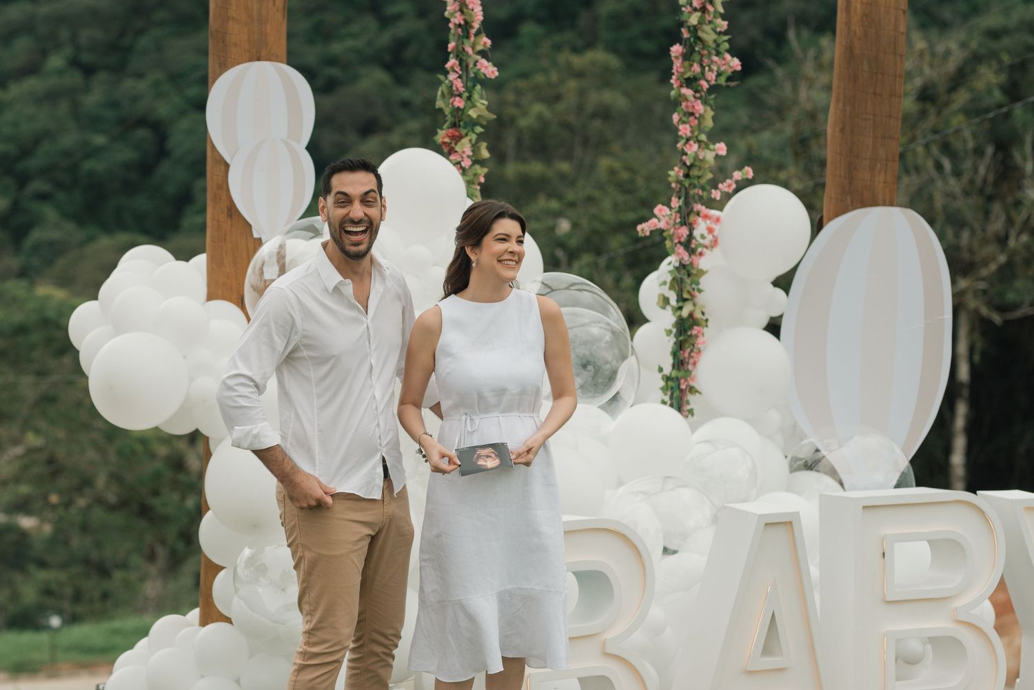 A baby shower celebration setup with white balloon decorations and floral accents under a wooden arch.