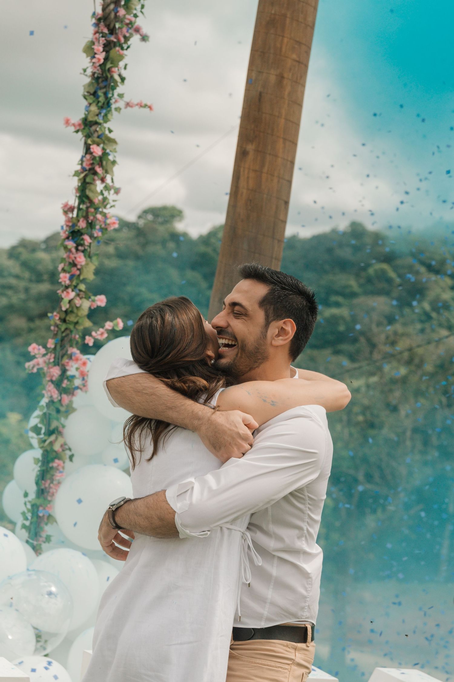 Couple embraces under wooden arch with floral decorations and pink flowers against mountain backdrop.